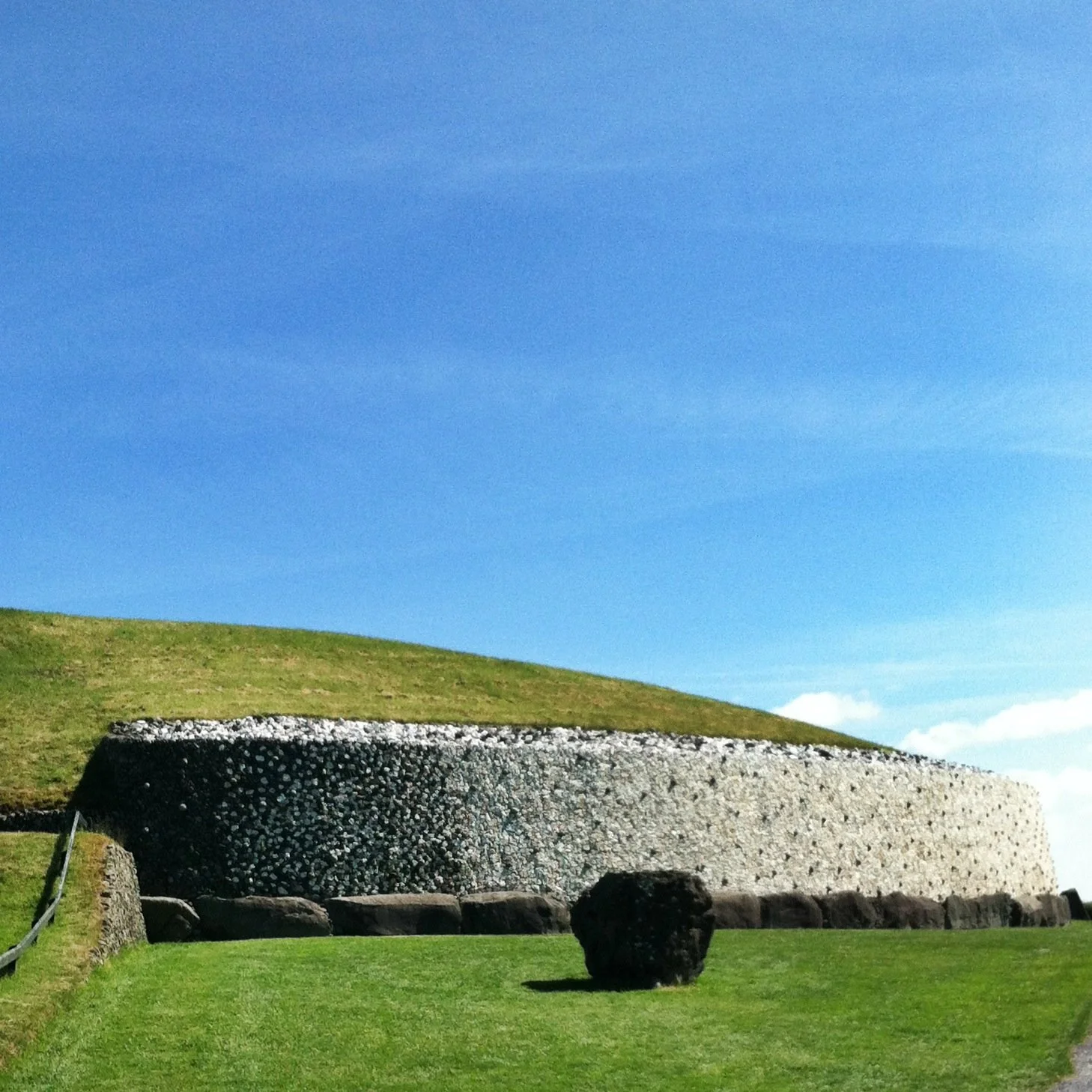 A large stone structure with a rough, pebble-textured surface is situated on a grassy landscape under a clear blue sky. There is a small black rock in the foreground and a hill behind the structure.
