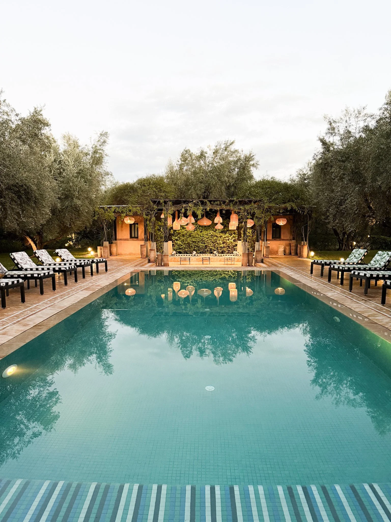 Infinity pool with black and white striped lounge chairs lining both sides, surrounded by lush green trees. In the background, a wooden stage with hanging lanterns and outdoor lighting, set for an event or performance, under an overcast sky.