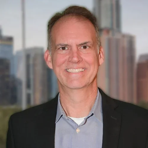 A man with short brown hair, wearing a black blazer and light blue collared shirt, smiling in front of a cityscape with tall buildings and a cloudy sky.