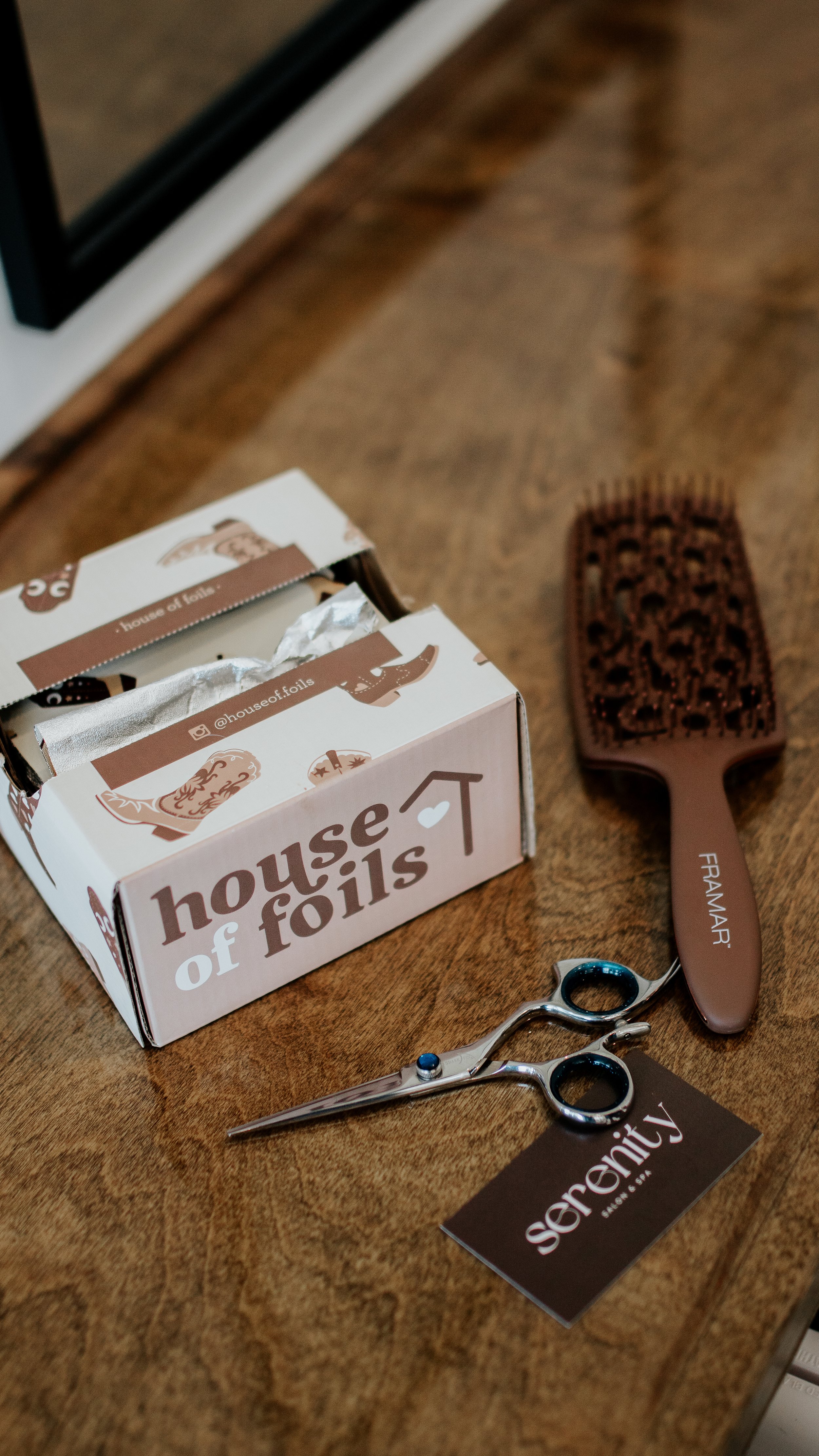 Box of hair foils, hairbrush, scissors, and business card on a wooden table.