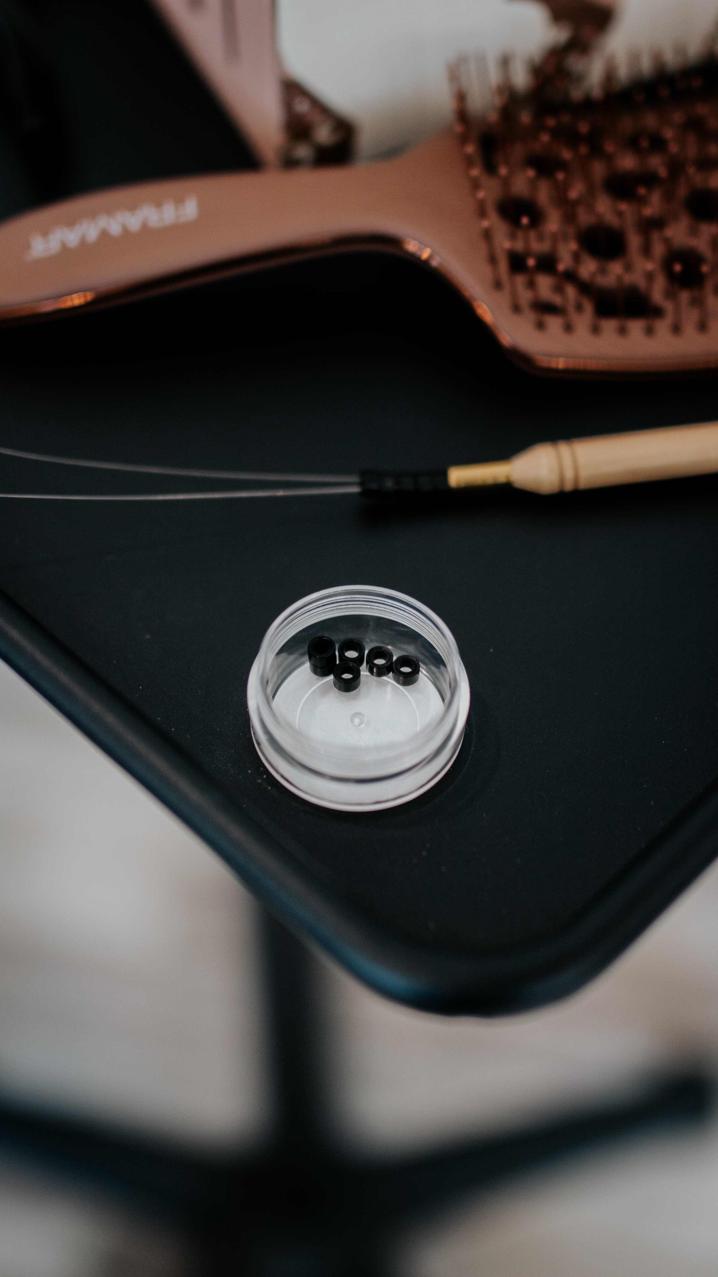 Close-up of hair extension micro rings in a small container on a table with a hairbrush and hair extension tool.