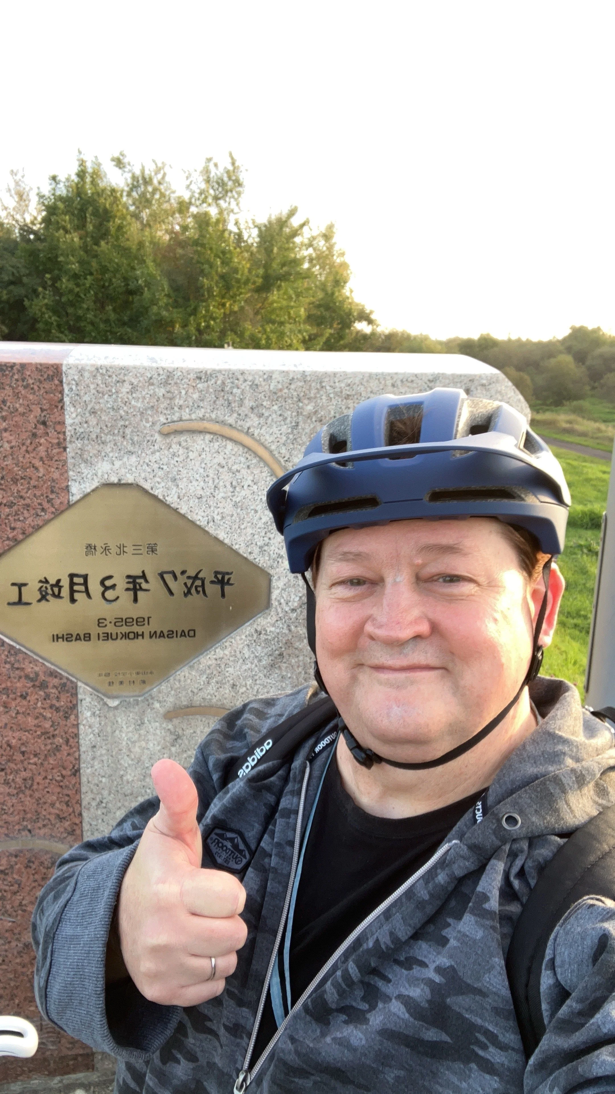 A man wearing a helmet and outdoor jacket giving a thumbs-up in front of a sign that reads 'Daishan Harbour District' in both Chinese and English, with trees and greenery in the background.
