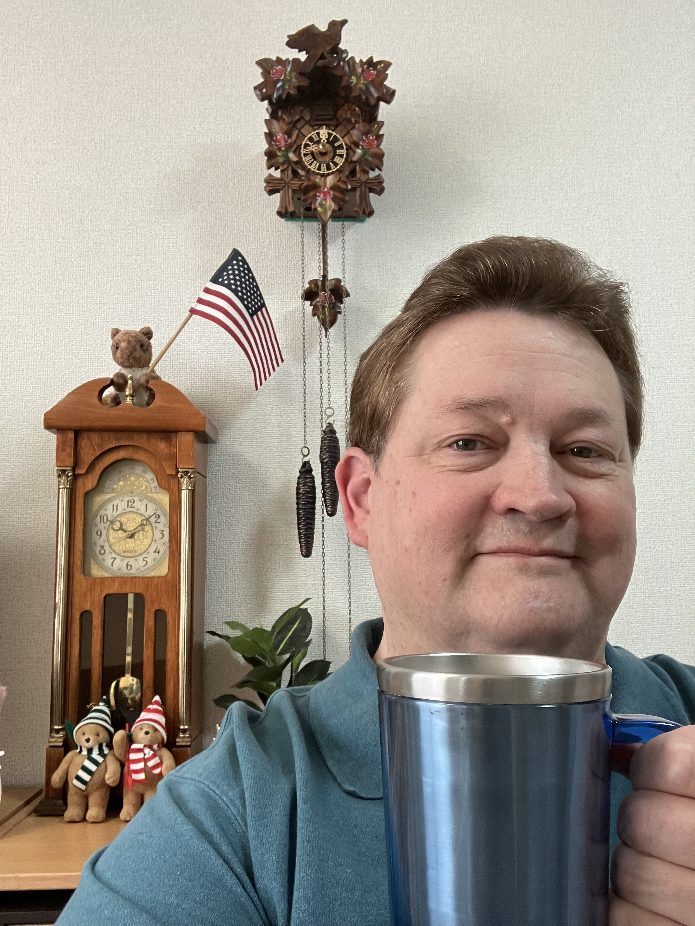 A man smiling and holding a blue and silver mug in front of a background with a wooden cuckoo clock, a small teddy bear with an American flag, two teddy bears with winter hats and scarves, and a houseplant.
