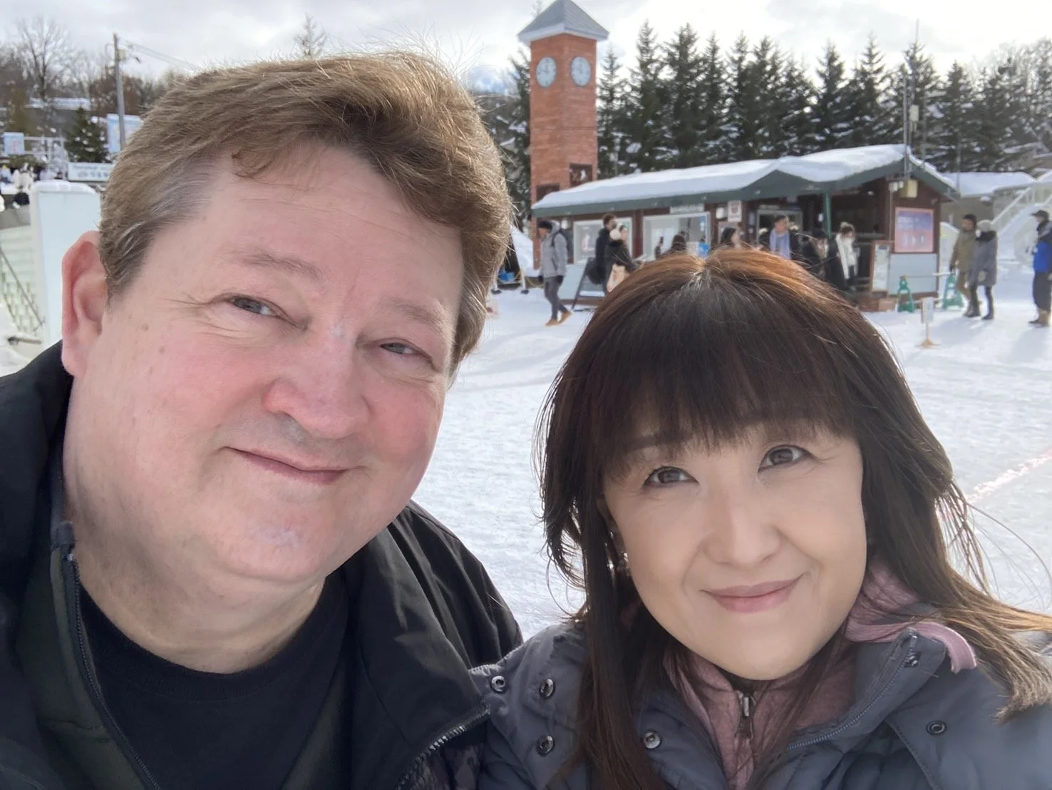 A man and a woman taking a selfie outdoors in a snowy area with a clock tower and a building with people in the background.