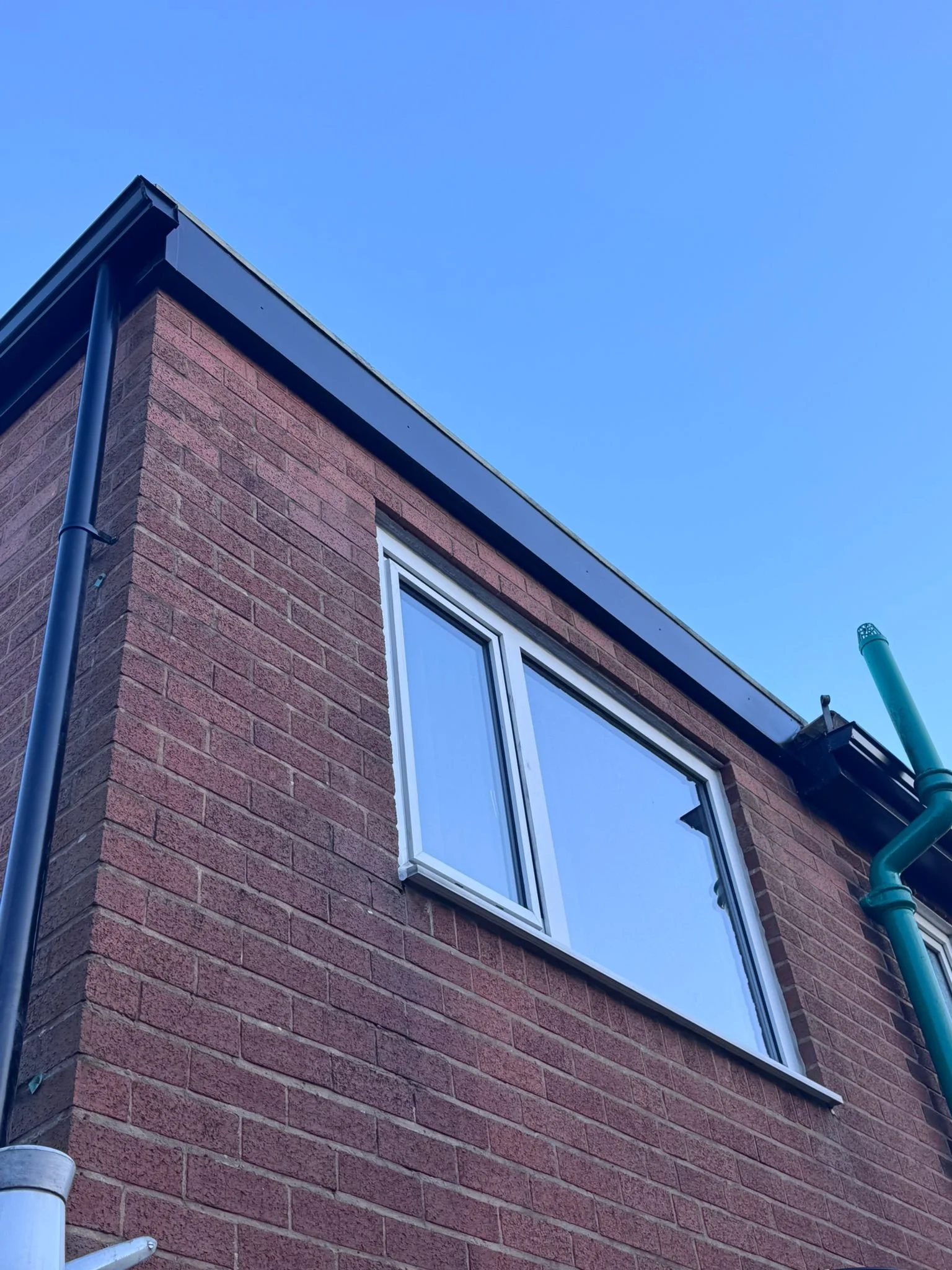 A corner of a brick building with a window, black gutters, and downspouts against a clear blue sky.