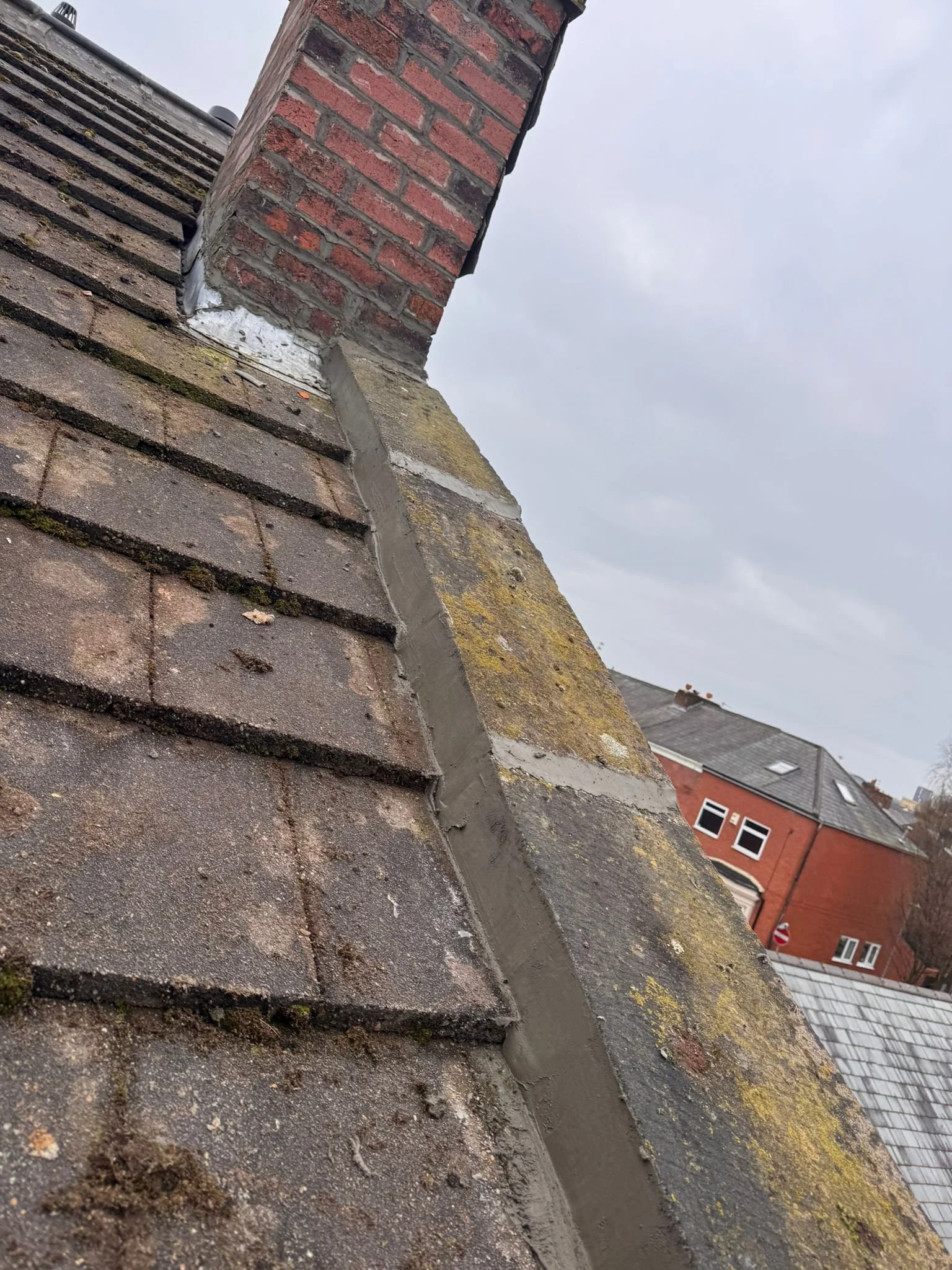 Close-up view of a rooftop with a chimney. The chimney is made of red bricks, surrounded by shingles that have moss growth and dirt on them. Background shows cloudy sky and neighboring buildings.