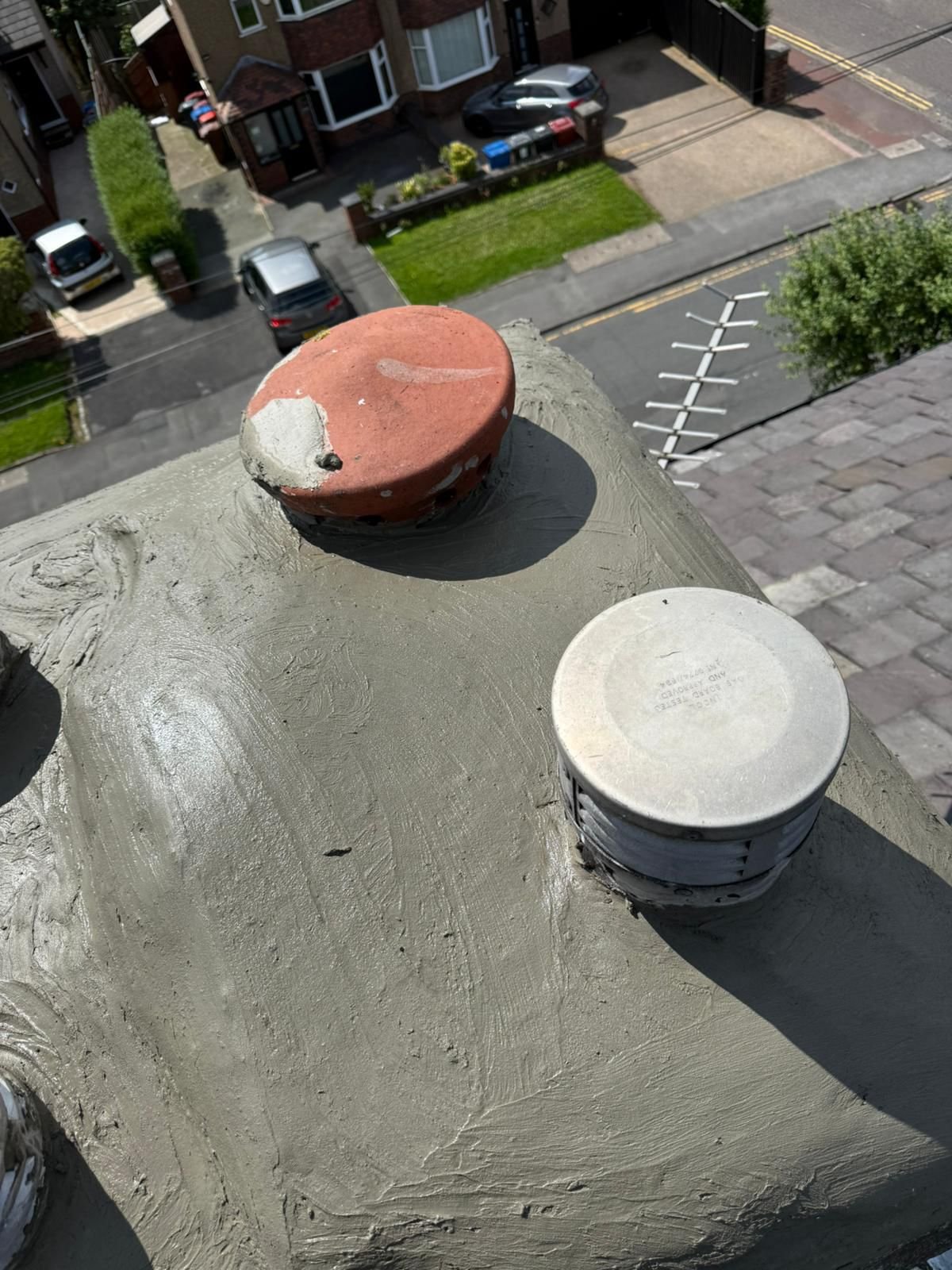 Close-up of a chimney with a newly applied layer of gray cement, showing two ventilation caps, one rusty red and one white, on top of the chimney in an urban residential area with parked cars, sidewalks, and greenery in the background.