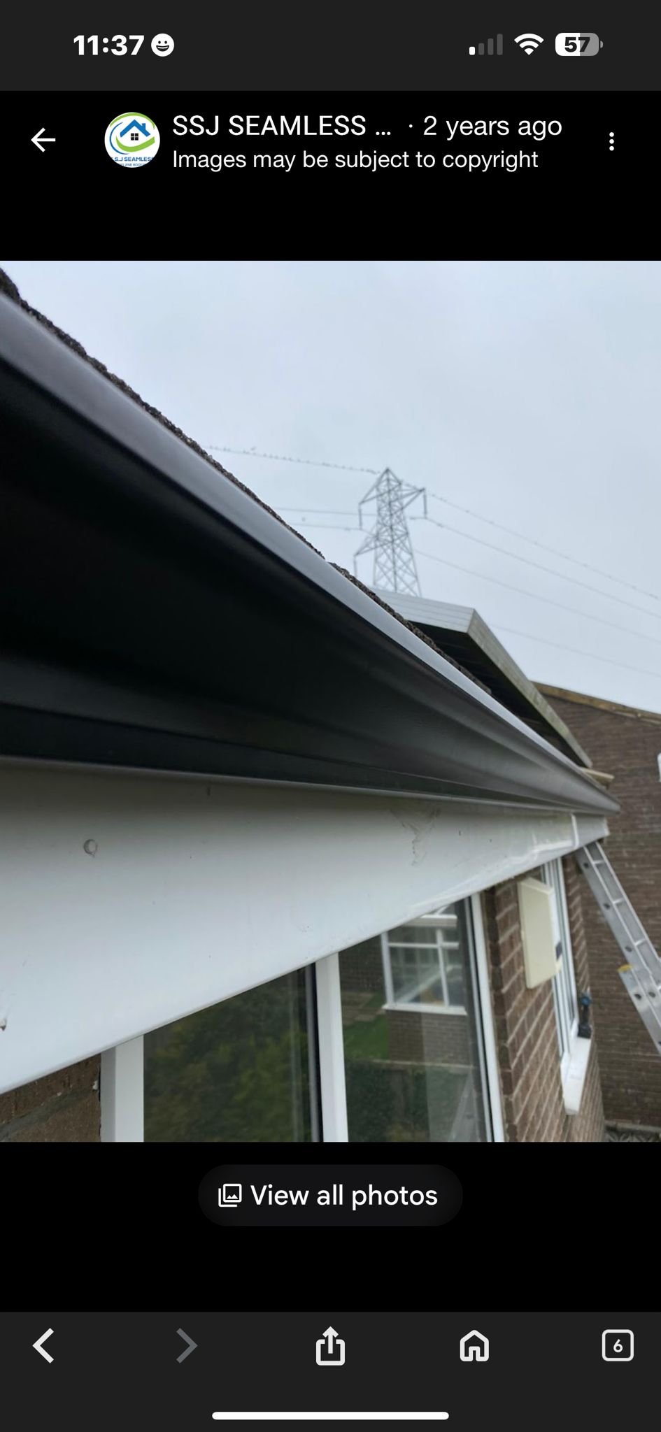 Close-up view of a house's gutter and roof with a ladder leaning against the side of the house, in cloudy weather. In the background, there is an electrical tower and power lines.