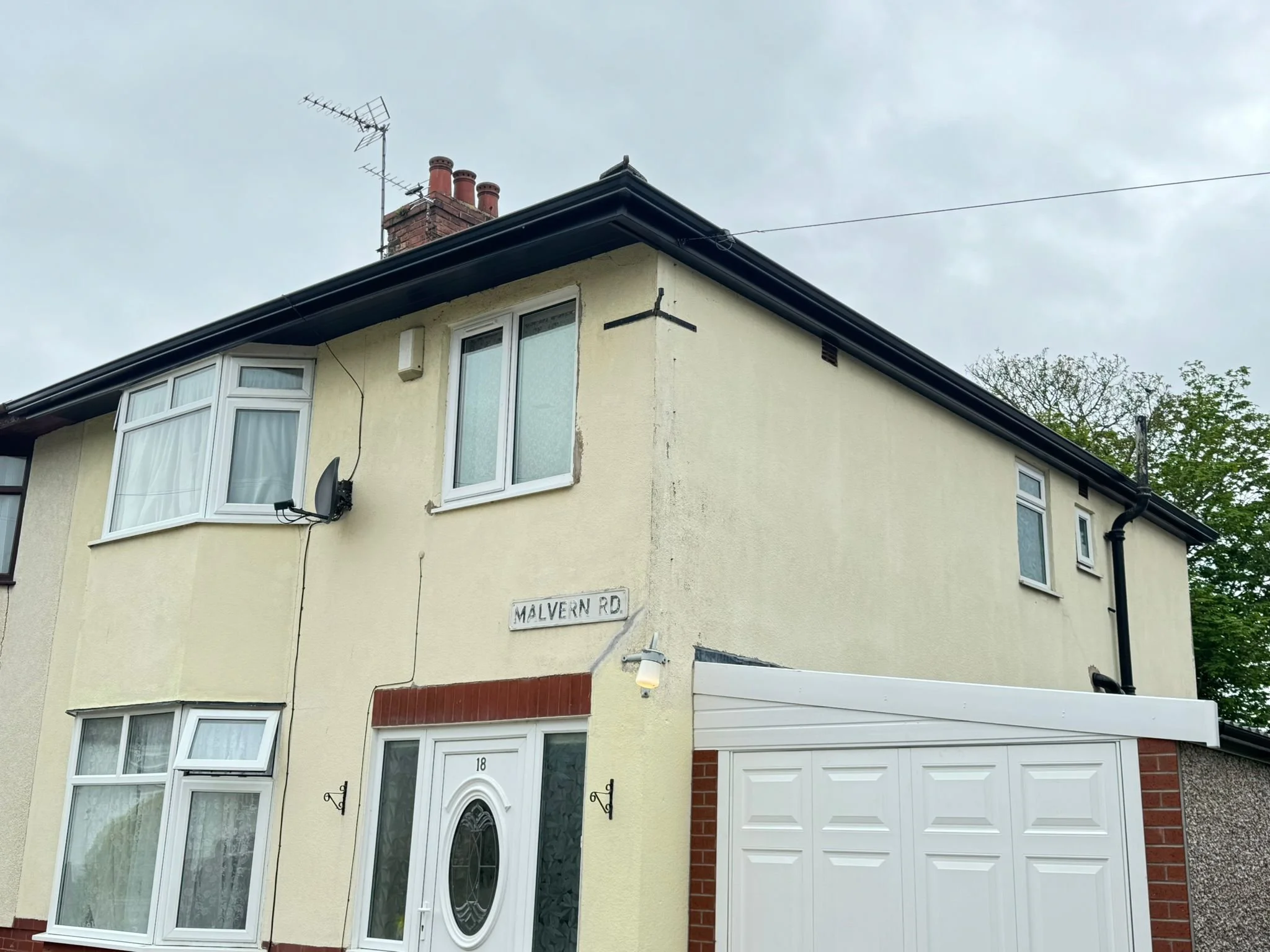 A yellow residential building with white-framed windows and a white front door, a garage with a white door, a satellite dish, and a street sign reading 'Malvern Rd.' under an overcast sky.