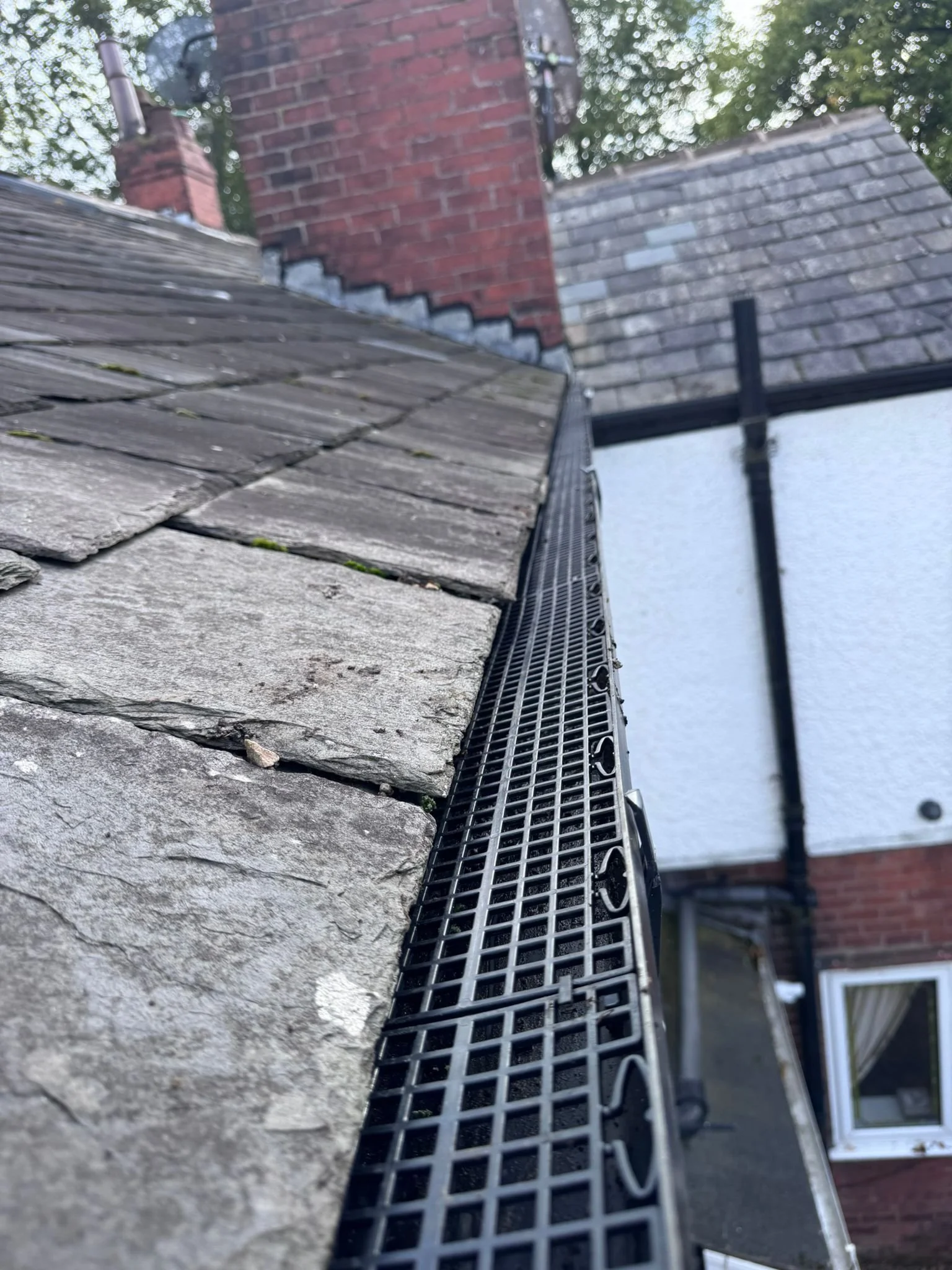 Close-up of a roof with gray shingles and a black gutter with a metal grate, on a building with brick chimney and white exterior wall, trees in the background.