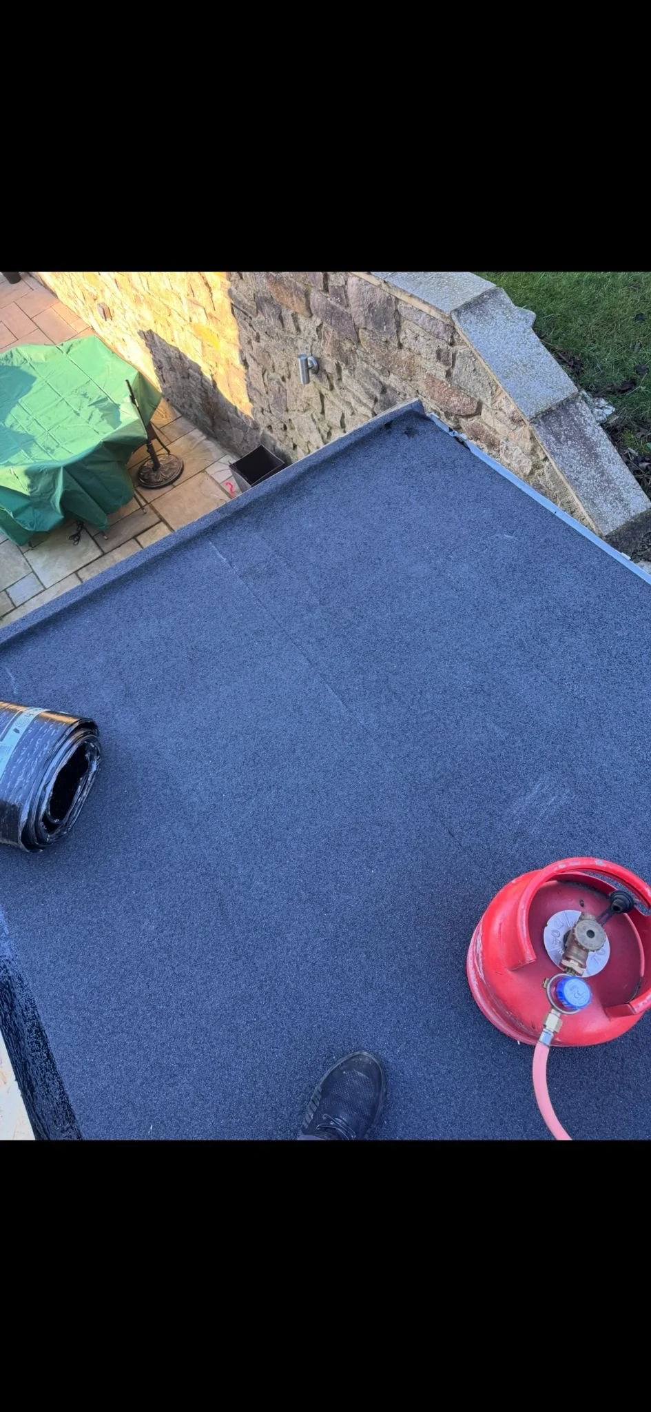 View from a rooftop showing a black rubber-like surface, a red propane tank with a pink hose, a roll of black roofing material, and a person's black shoe. Below is an outdoor patio area with a green covered table, a lamp, a small black container, and