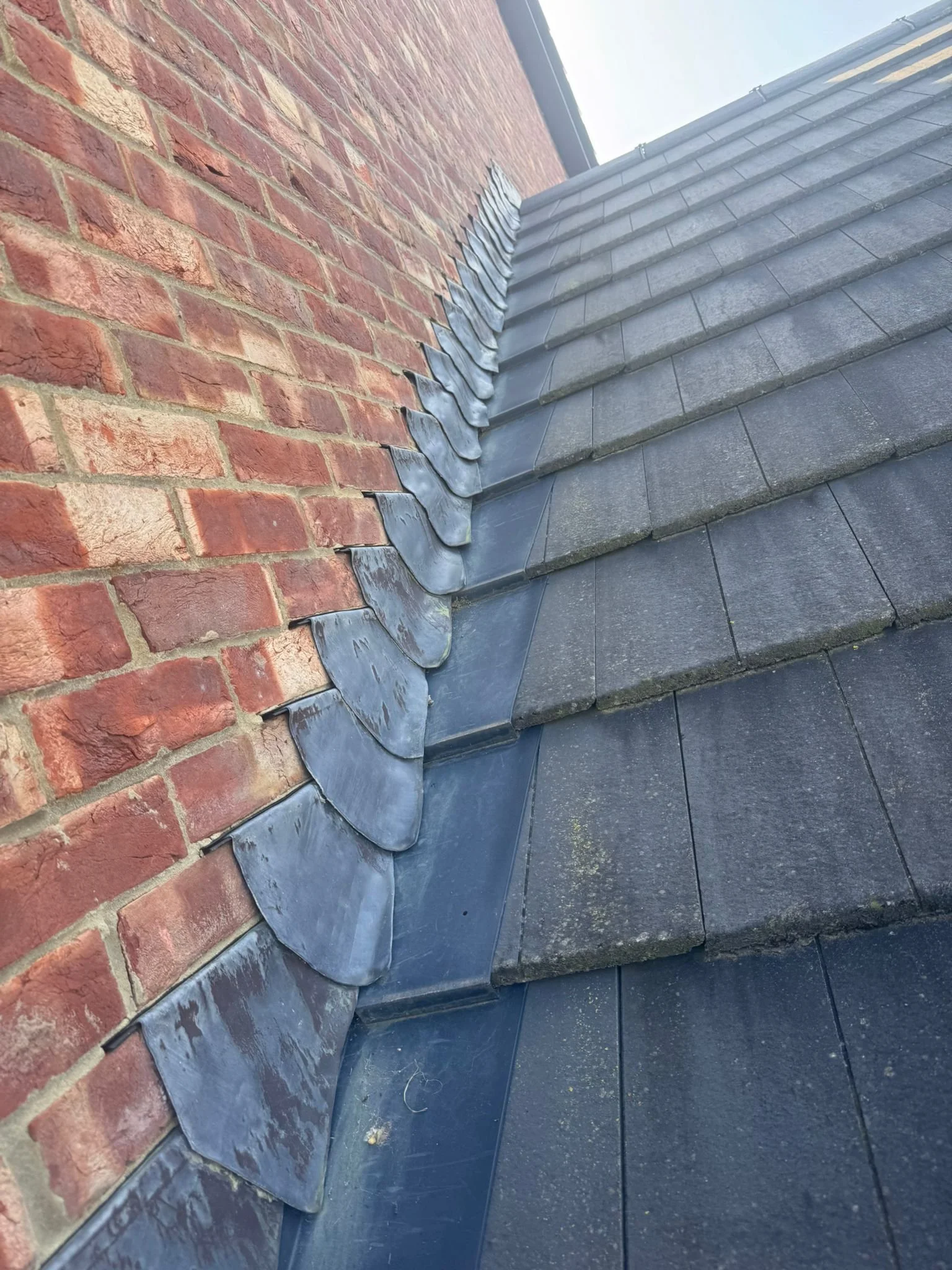 Close-up of a roof with dark grey tiles and a red brick wall showing a row of metal flashing along the edge of the roof.