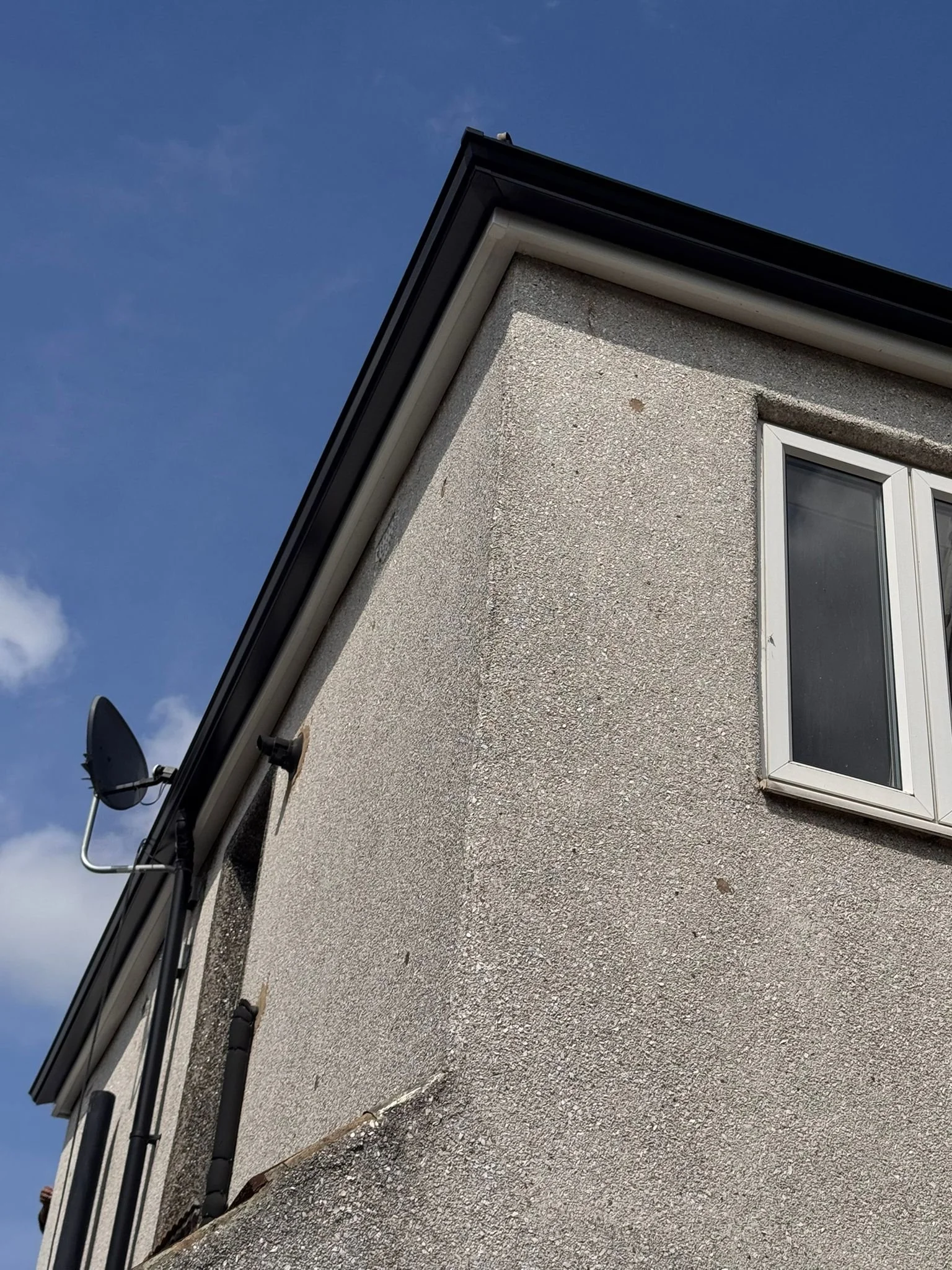 Part of a beige stucco house with a white-framed window in the bottom right corner, black roof shingles, and a satellite dish mounted on the left side. A blue sky with a few clouds is in the background.