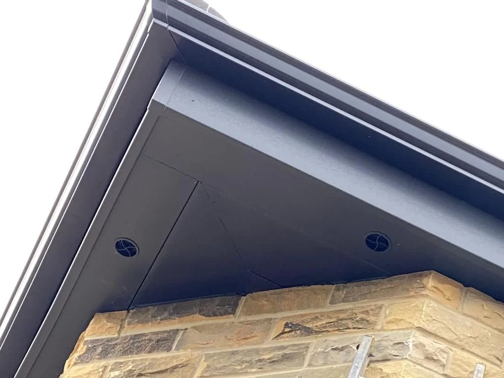 Close-up of the corner of a building showing a gray metal roof over a stone wall, with ventilation holes in the roof soffit.