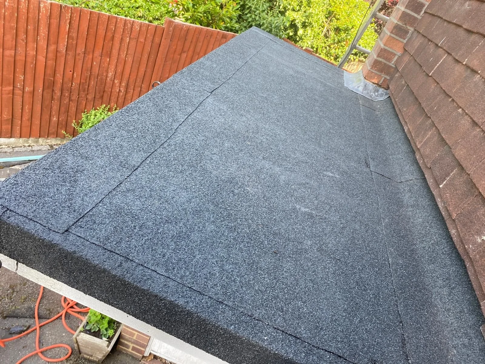 Close-up of a roof with black asphalt shingles, showing the edges and seams, with a brick chimney and a wooden fence in the background.