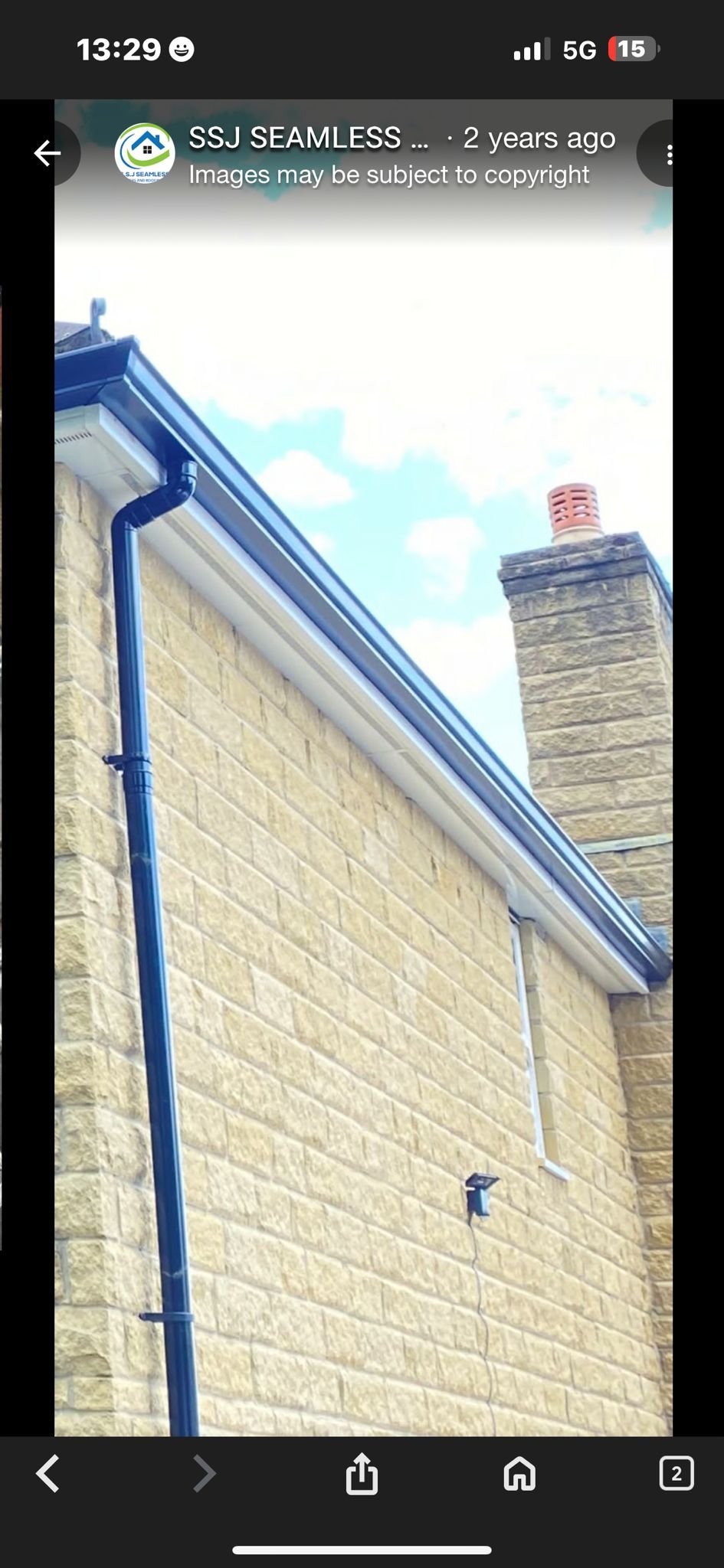 Close-up of a yellow brick house wall with a black gutter, a white downspout, a small security camera, and a chimney with a vent on the roof under a partly cloudy sky.