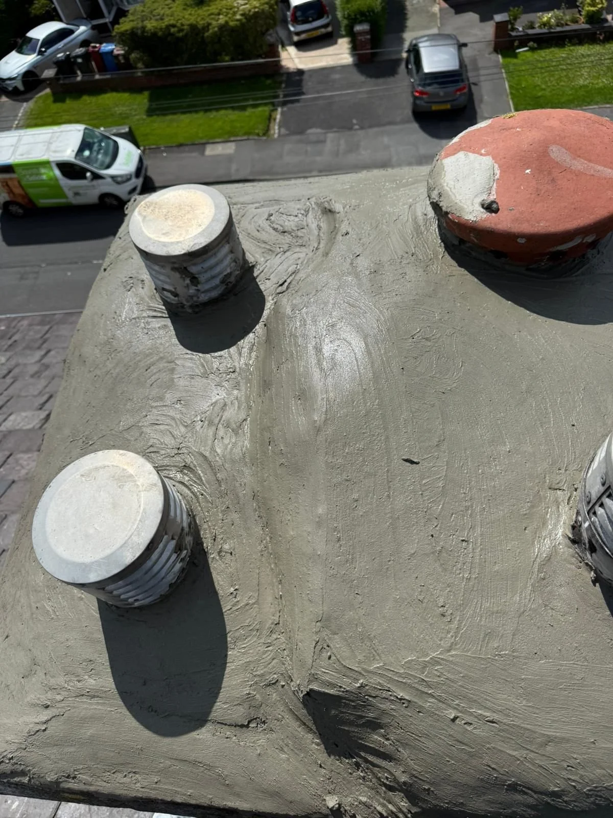 Close-up of a chimney top under repair, with fresh cement and some chimney caps visible, overlooking a residential street with parked cars and green lawns.