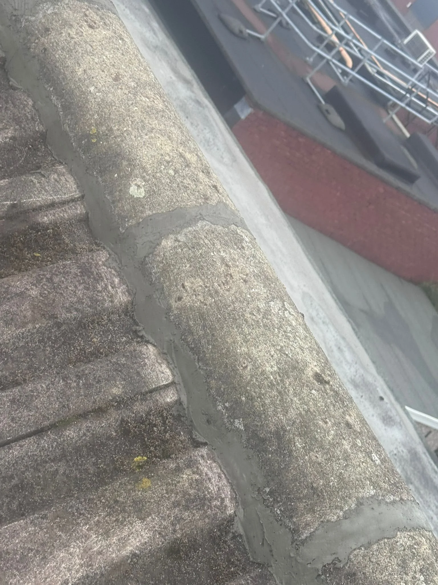 Close-up of a section of concrete roof with visible mortar joints and part of a building with debris and structure in the background.