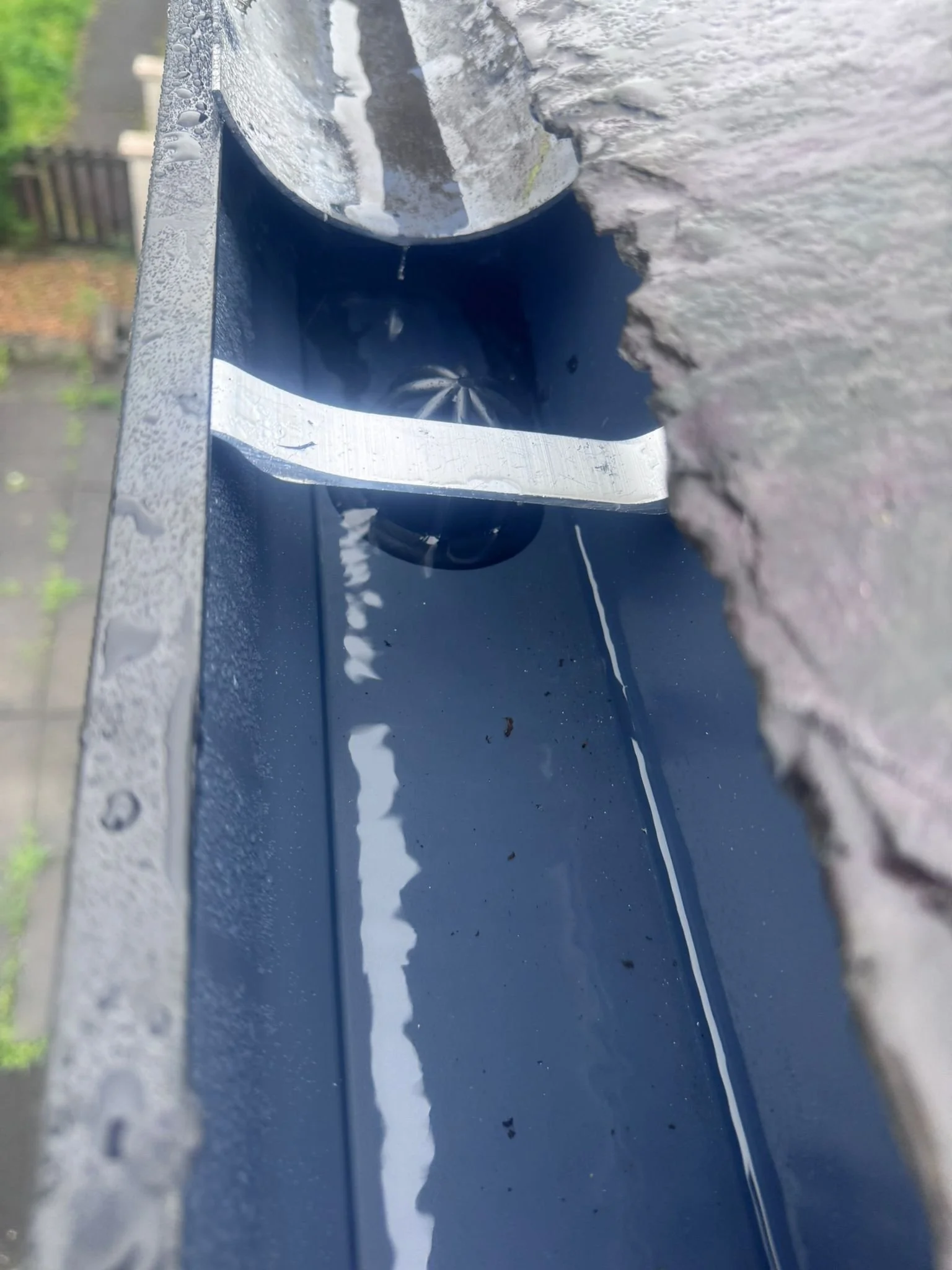 Close-up of a rain gutter with water inside, showing raindrops on the metal surface and a reflection of the sky and trees in the water.