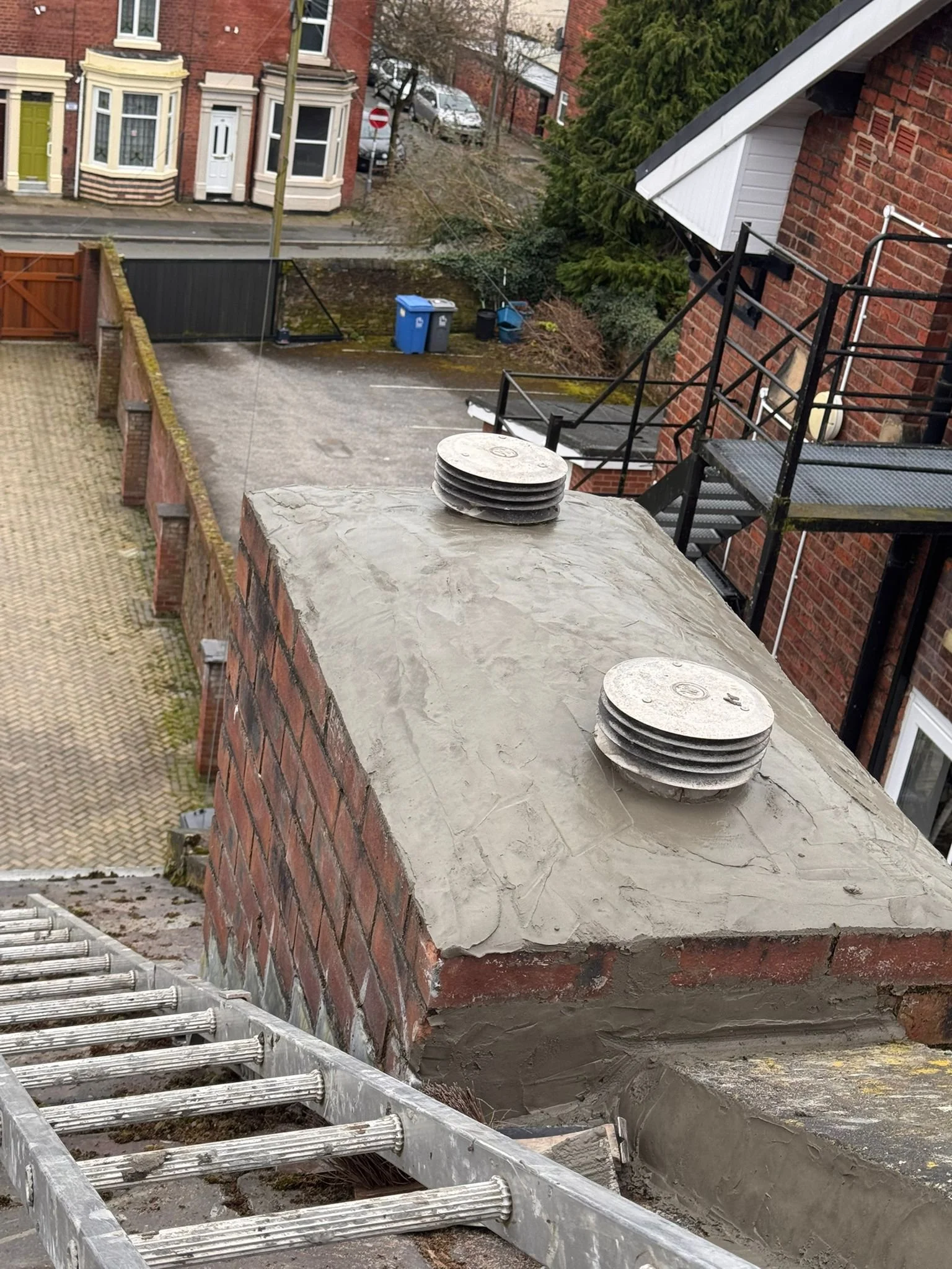 View from balcony showing brick building, metal staircase, tools, and a ladder, with residential street and parked cars in background.