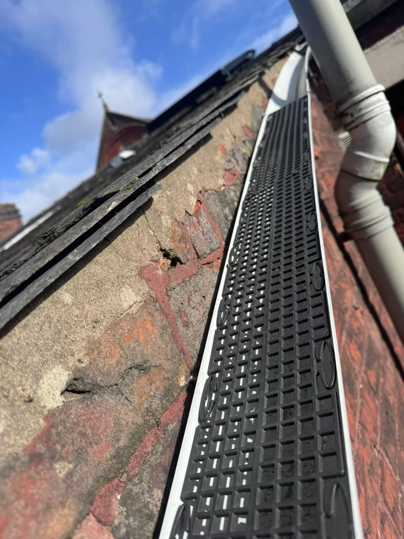 Close-up of a black drainage grate on a brick sidewalk, with a white pipe running alongside and a building with a brick wall and a tower in the background.