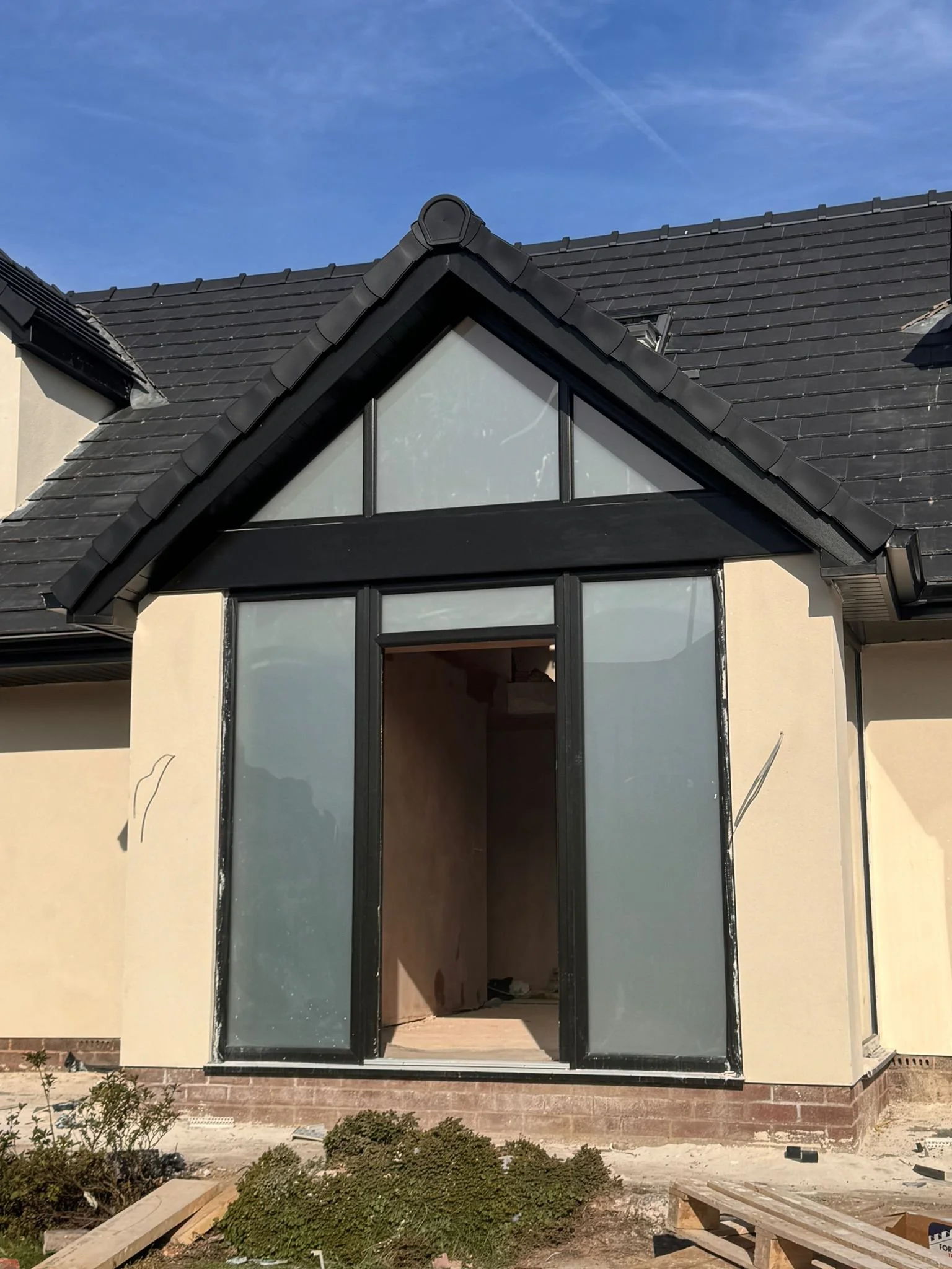 Front view of a house under construction with black window frames, a partially installed glass door, and a black tiled roof, with construction materials and plants in the foreground.