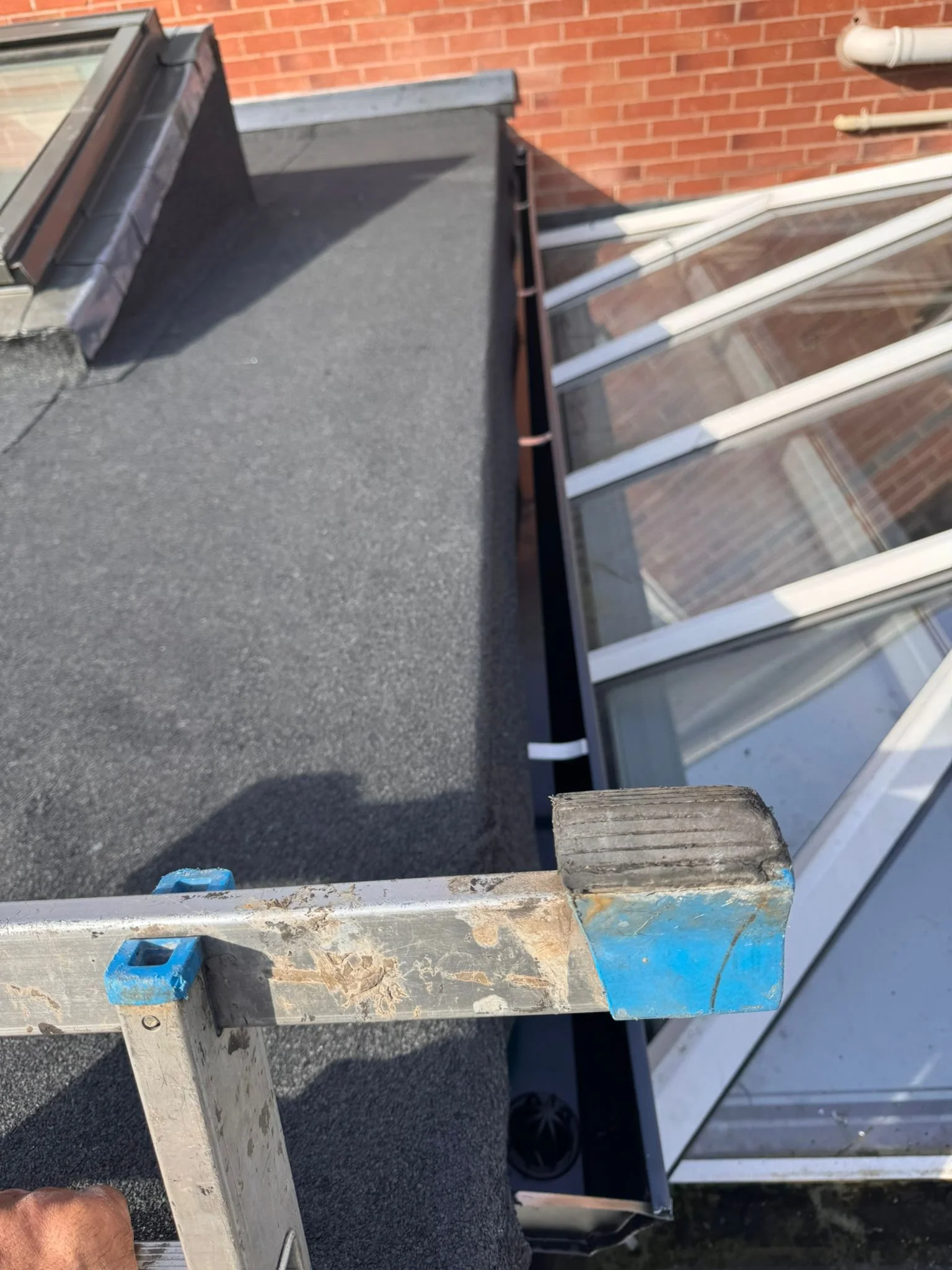 View of a rooftop with a flat black surface, part of a brick building wall in the background, and a metal railing in the foreground. There are white metal bars and glass panels, possibly a sunroom or enclosed balcony. A person's hand and a blue objec