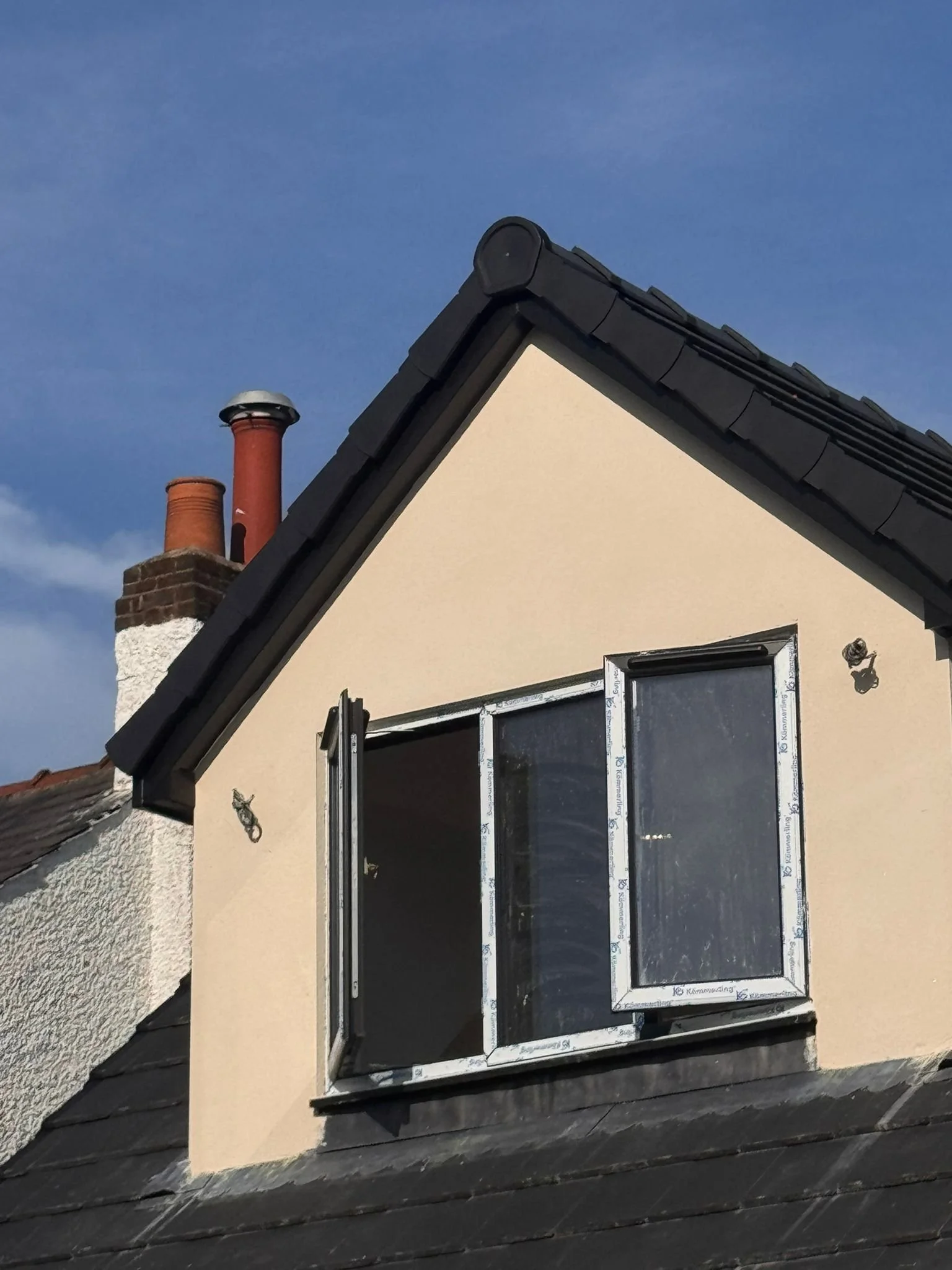 The image shows the upper part of a house with a black tiled roof, a cream-colored wall, and a double dormer window with black trim. There are two brick chimneys in the background against a blue sky.