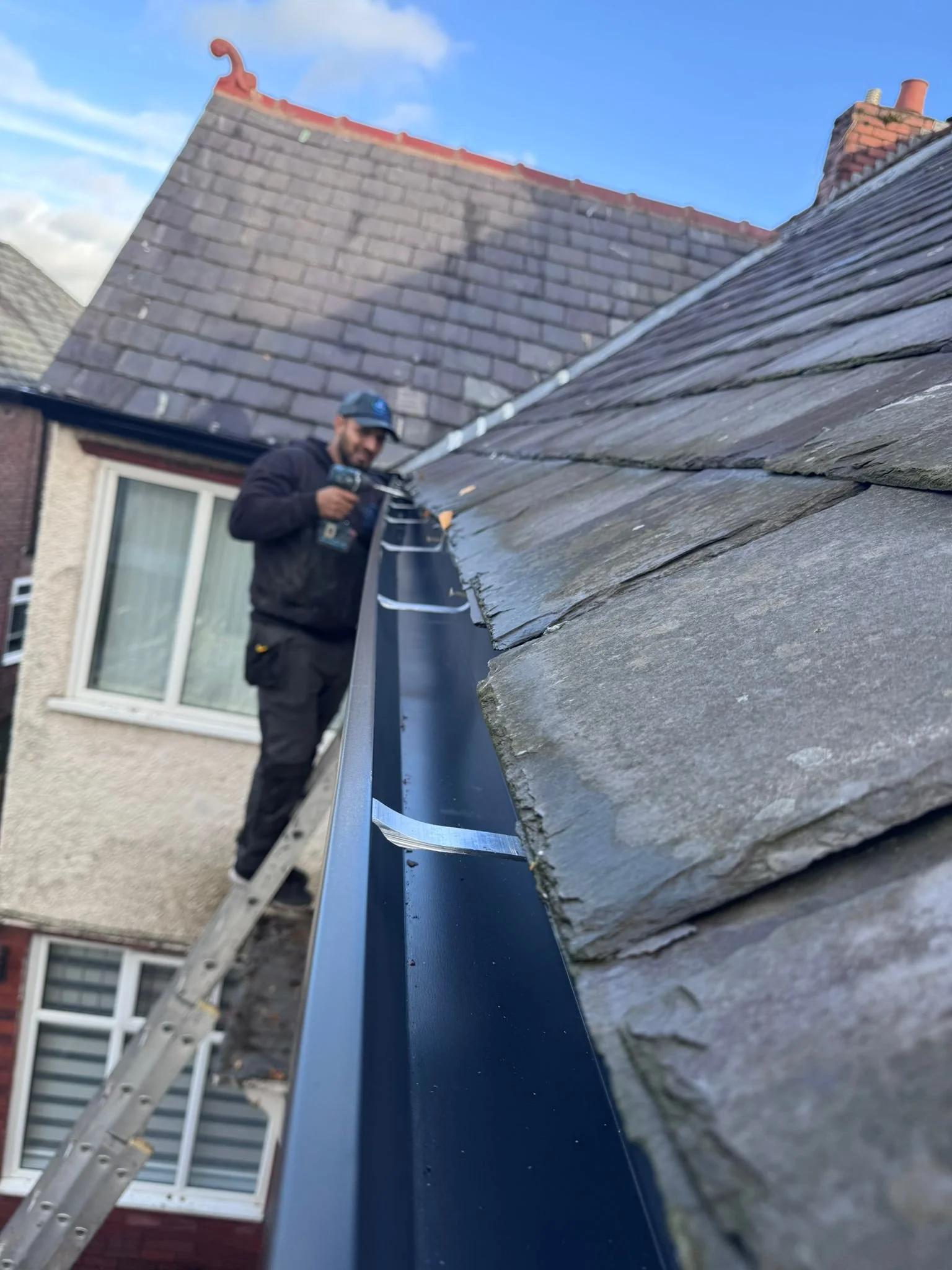 A person working on a roof with a ladder leaning against the house, installing or repairing a blue gutter, with houses and a blue sky in the background.
