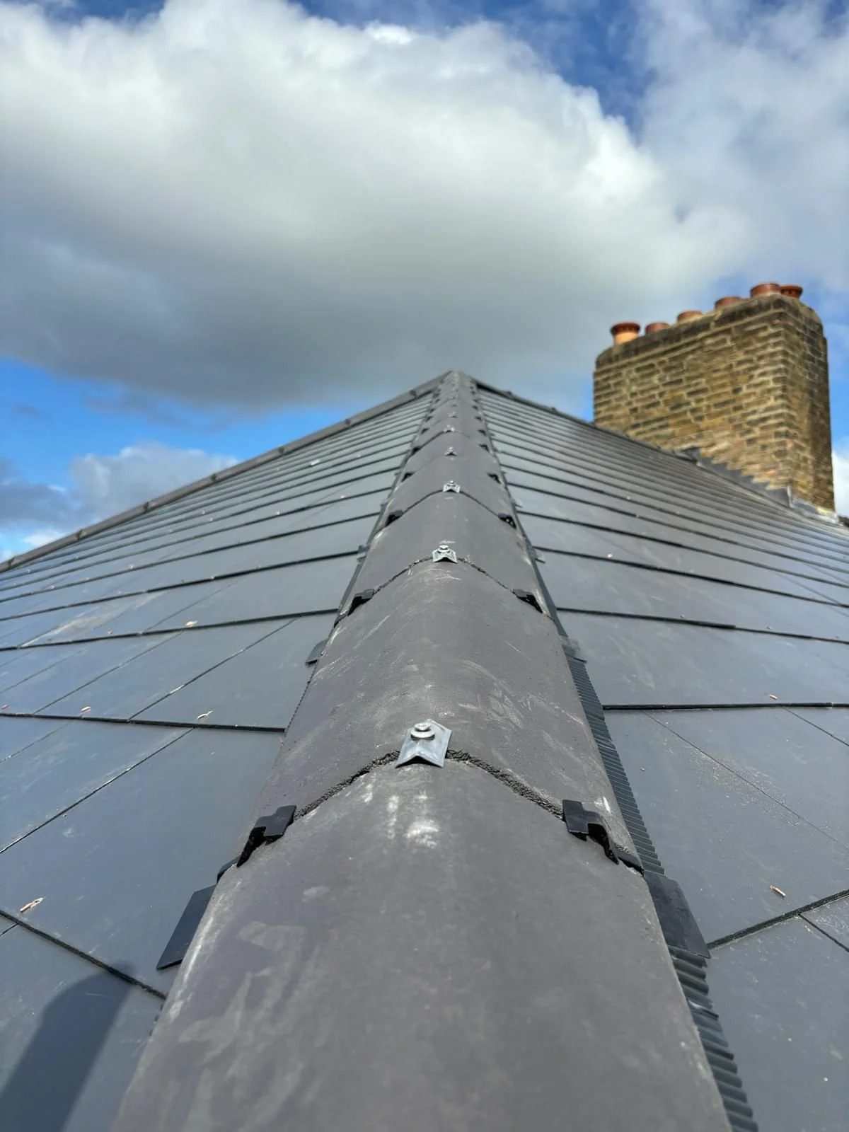 Looking up a sloped metal roof with a brick chimney and a cloudy sky in the background.