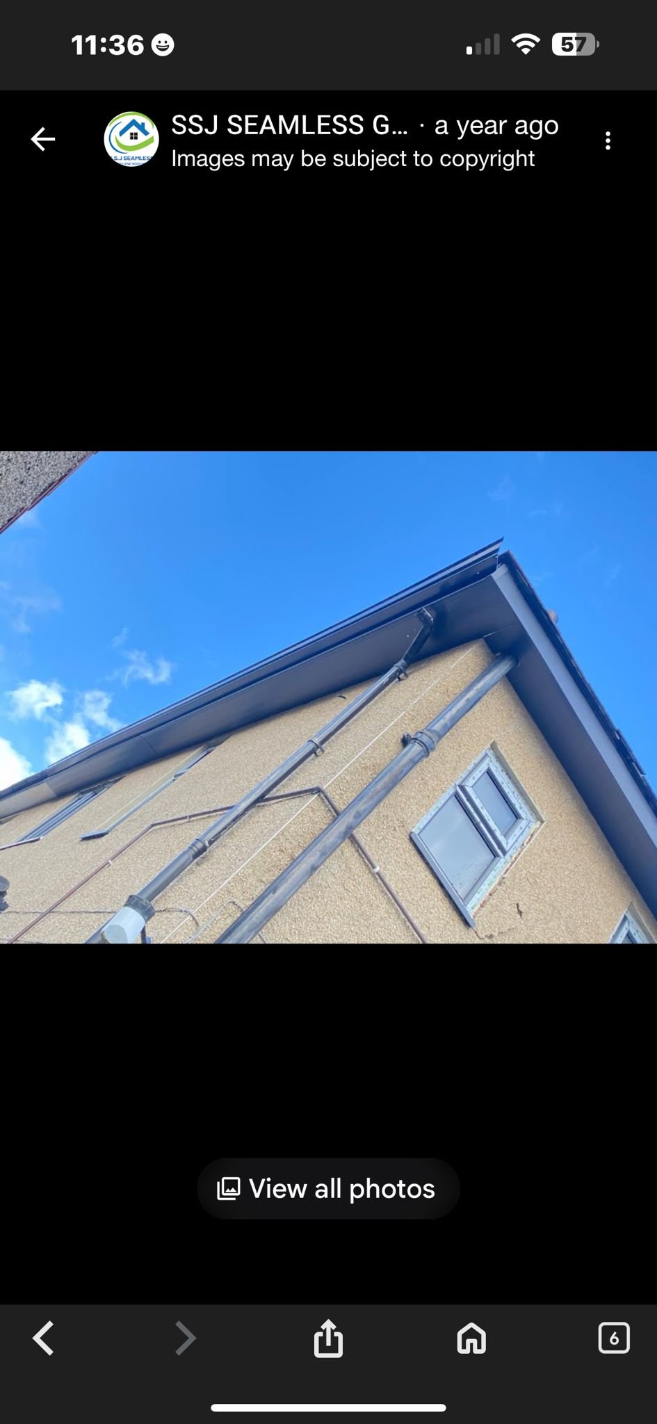 Photo of the upper corner of a residential building with beige textured walls, silver gutters, and metal pipes against a blue sky with some clouds.