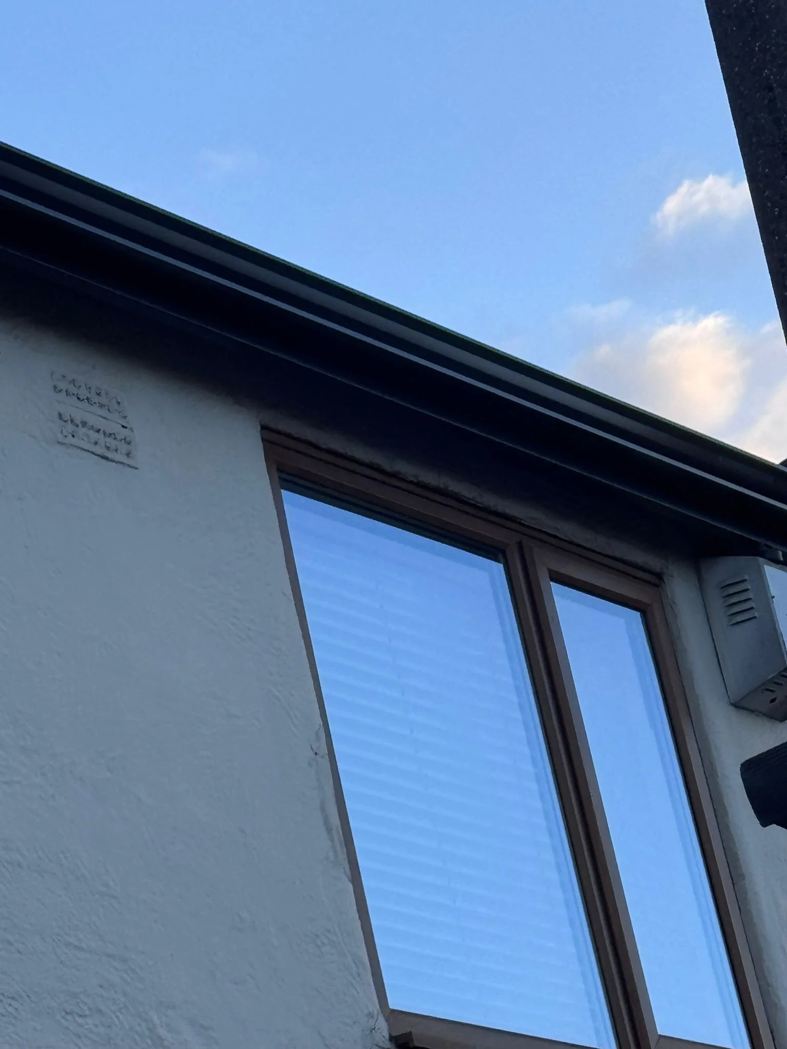 View of blue sky with some clouds through a window, part of a building's roof and wall are also visible.
