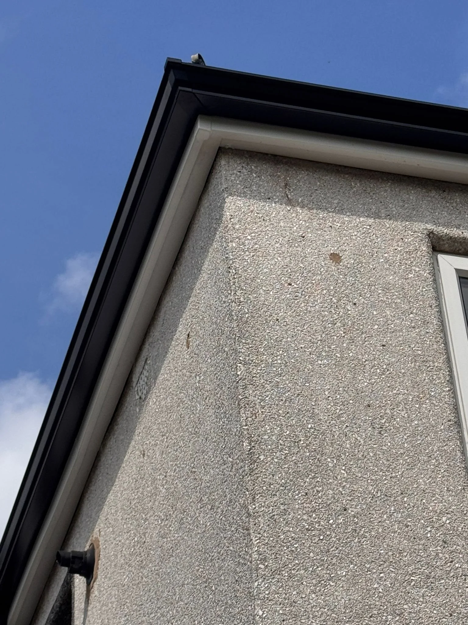 Close-up of a house exterior corner showing textured beige stucco wall, white trim, black roof edge, small white vent, and part of a window with a white frame. Blue sky with some clouds in the background.