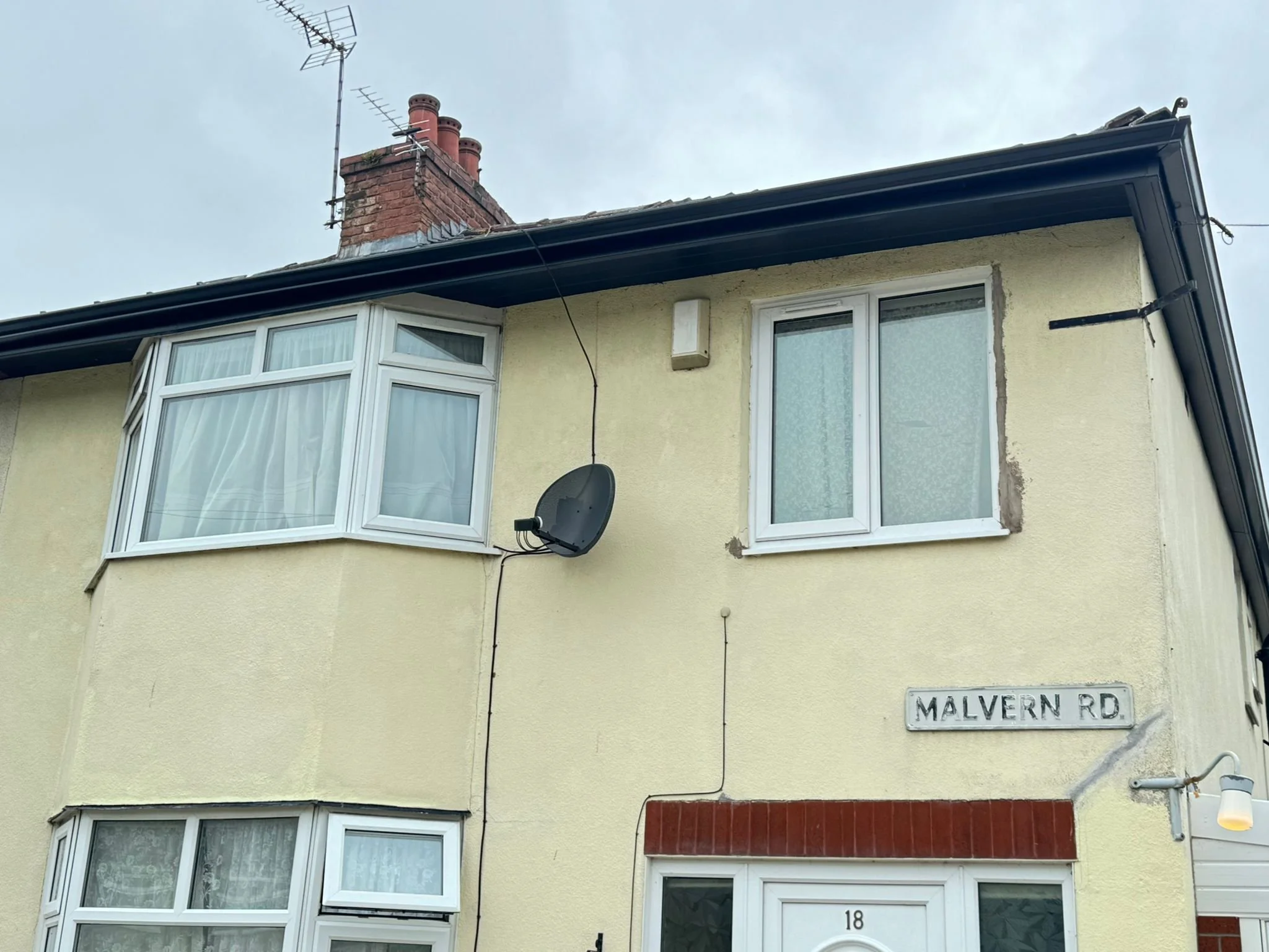 A two-story yellow house with white window frames and bay windows, a red brick chimney, and a satellite dish, located on Malvern Road.