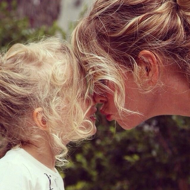 A woman and a young girl with curly blonde hair are touching foreheads outdoors, with their eyes closed, in a tender moment.