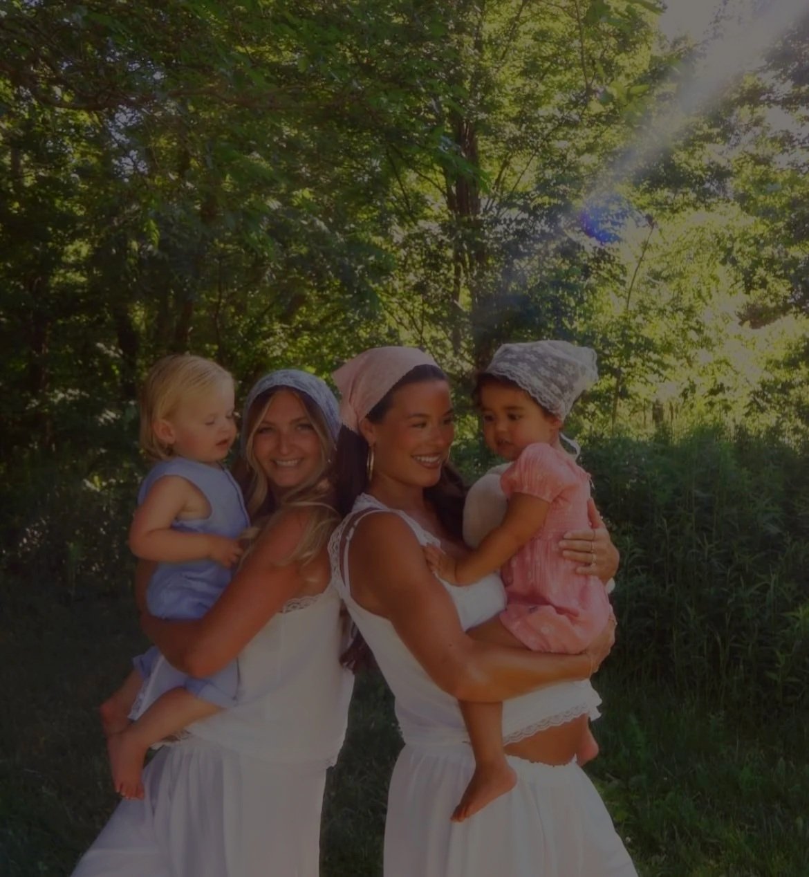 Two women in white dresses holding children in a forest with sunlight streaming through trees.