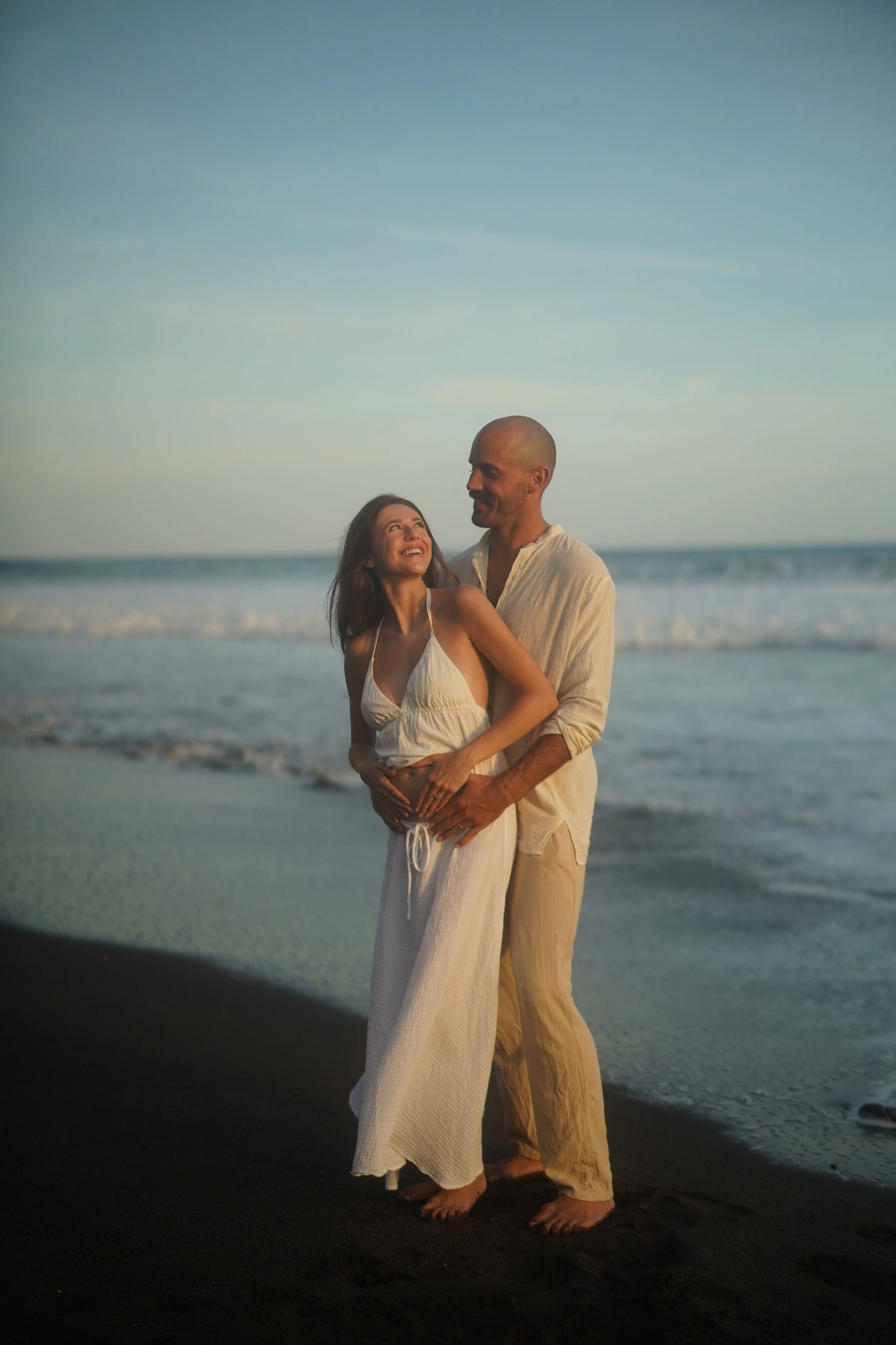 A couple standing on the beach, holding each other and smiling, with the ocean waves in the background during sunset.