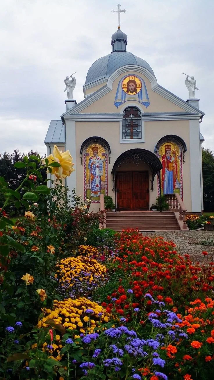 A small church with religious icons on the front, topped with a blue dome and a cross, surrounded by colorful flowers.