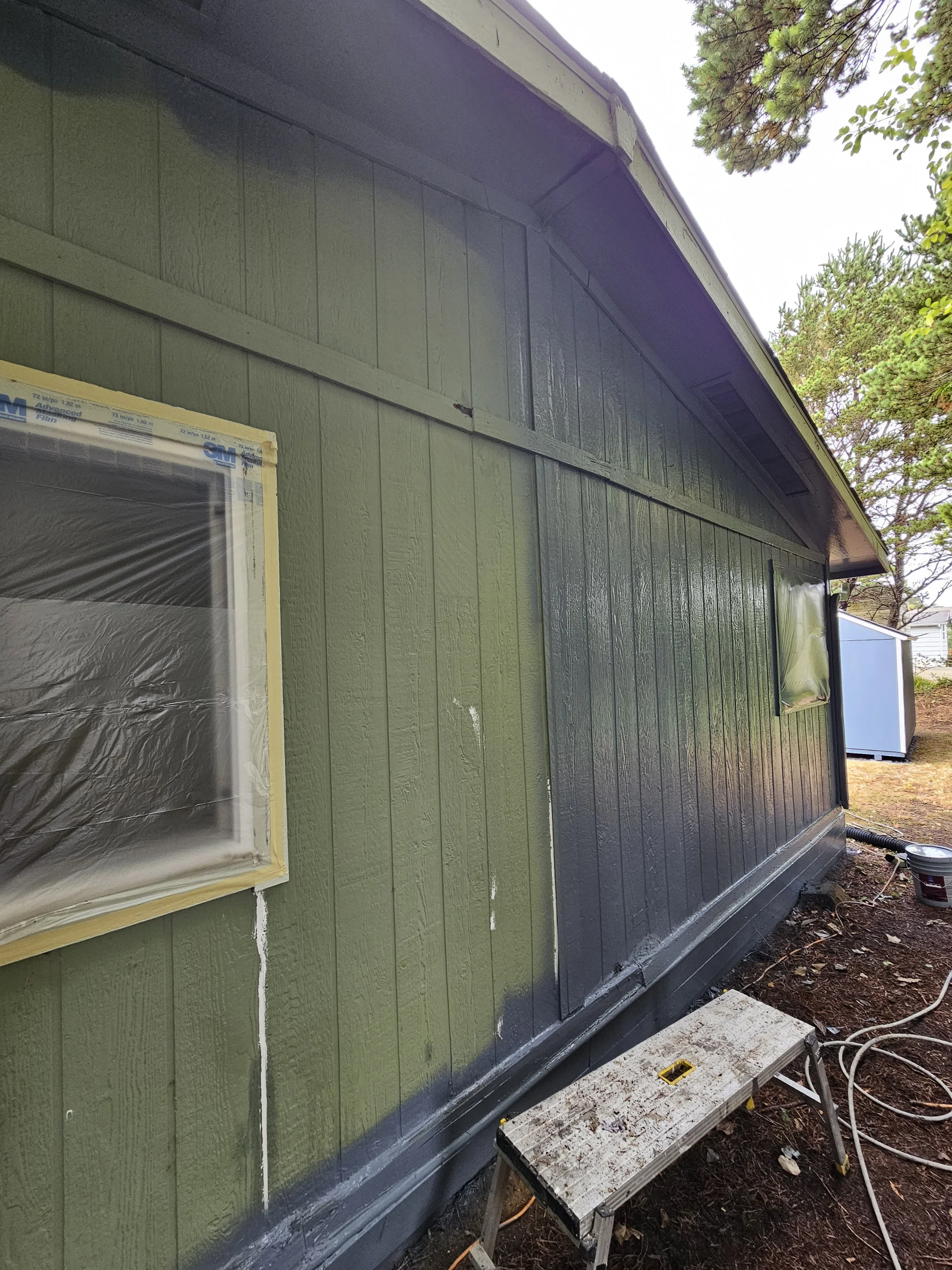 Side of a house being painted green, with a window covered in plastic, a small step stool, and construction tools on the ground.