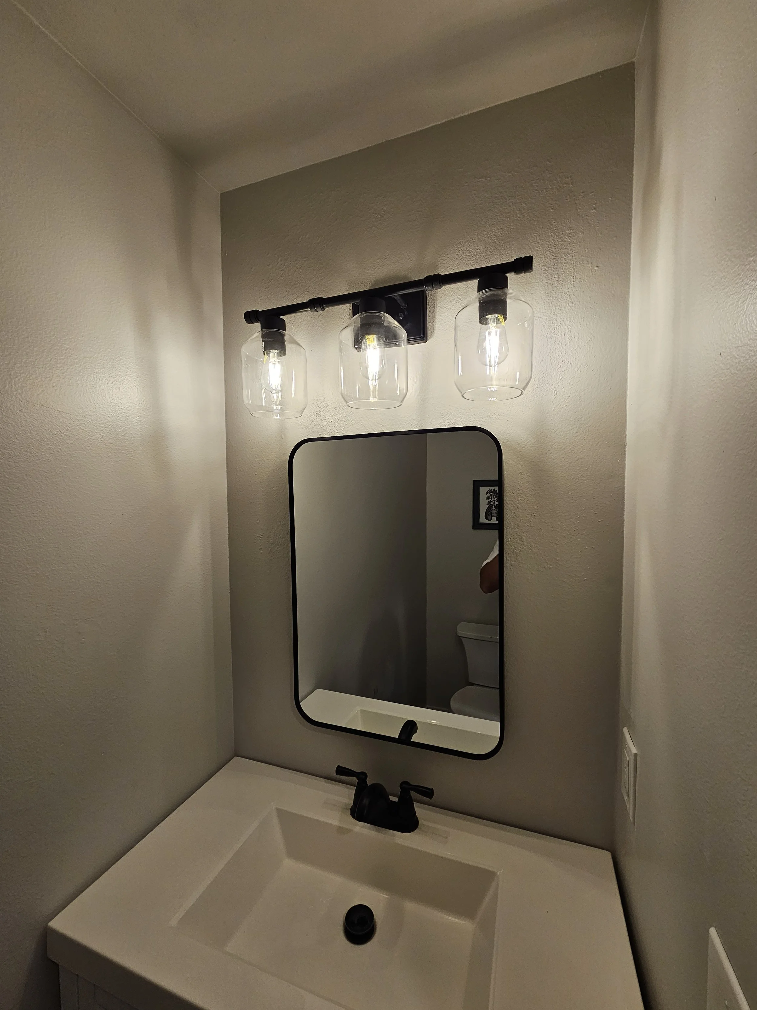 A bathroom vanity with a white sink, black faucet, a rectangular mirror with rounded edges, and a three-light vanity fixture with clear glass shades and exposed bulbs mounted above the mirror.
