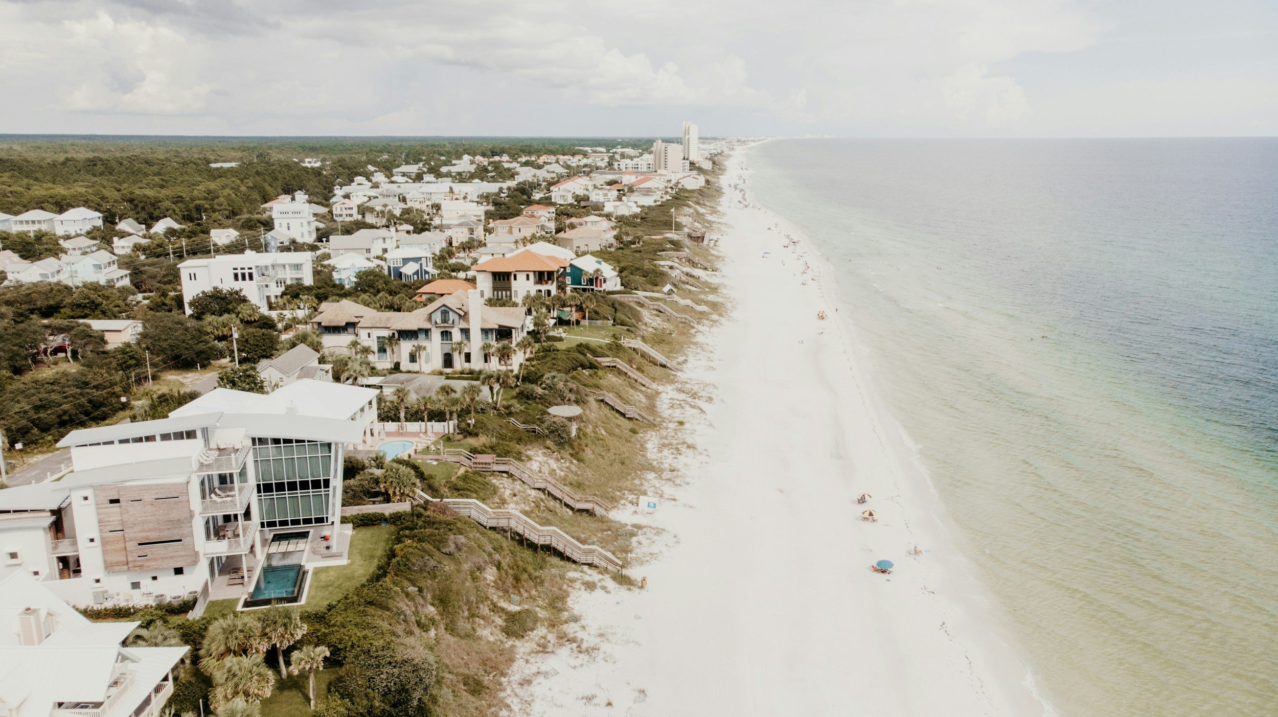 Aerial view of a beachfront neighborhood with houses along the coast and a sandy beach stretching out to the horizon on a partly cloudy day.