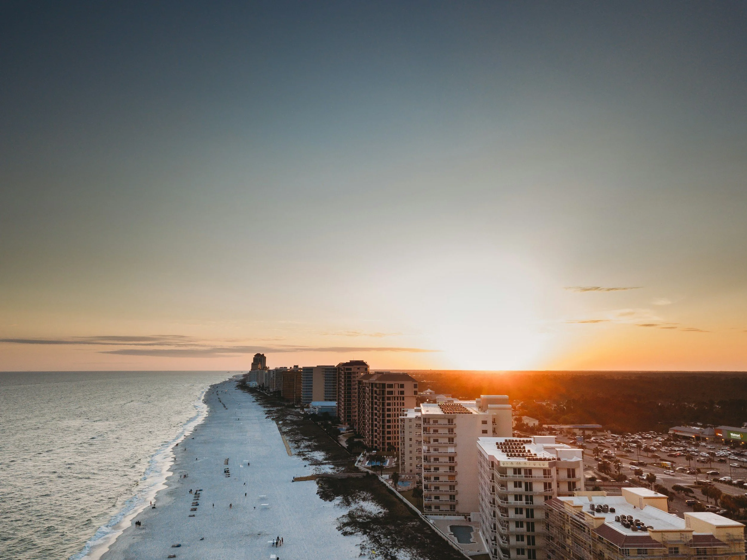 Aerial view of a beachside city during sunset with high-rise buildings along the shoreline and a parking lot nearby.