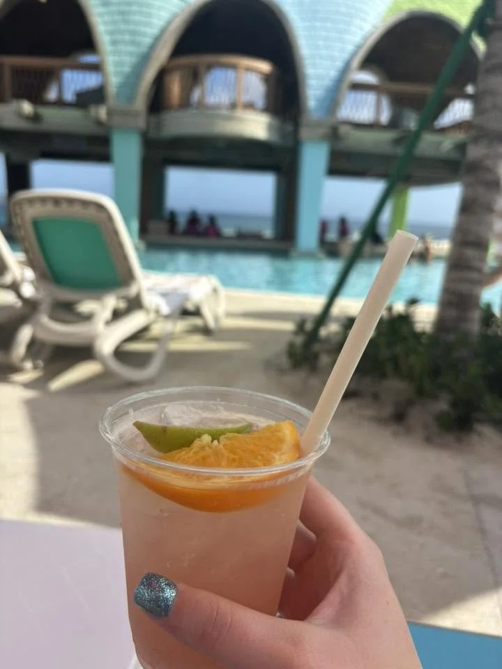 Hand holding a refreshing drink with orange and lime slices on a beachside patio with lounge chairs, a pool, and a blue ocean in the background.