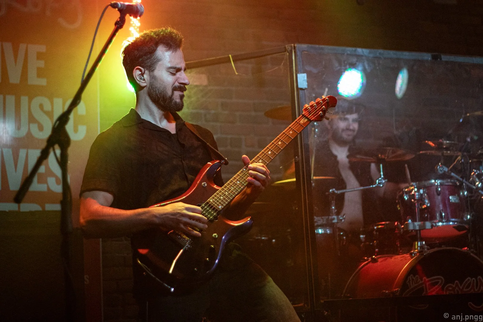 A man playing an electric guitar on stage with yellow and red stage lighting, a microscope and a brick wall in the background, and a drummer behind a transparent divider.