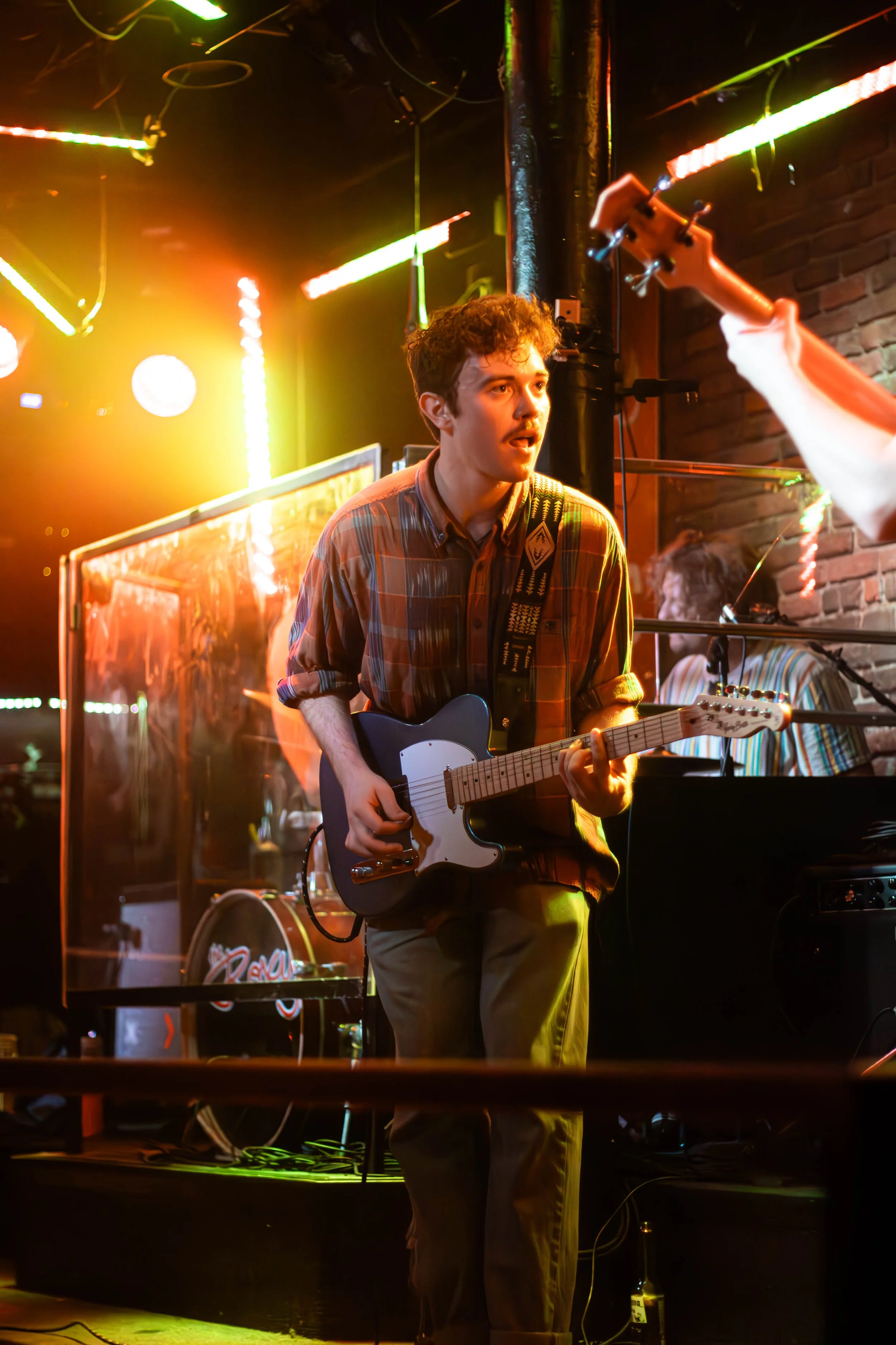 Young man playing an electric guitar on stage under colorful lighting at a music venue.