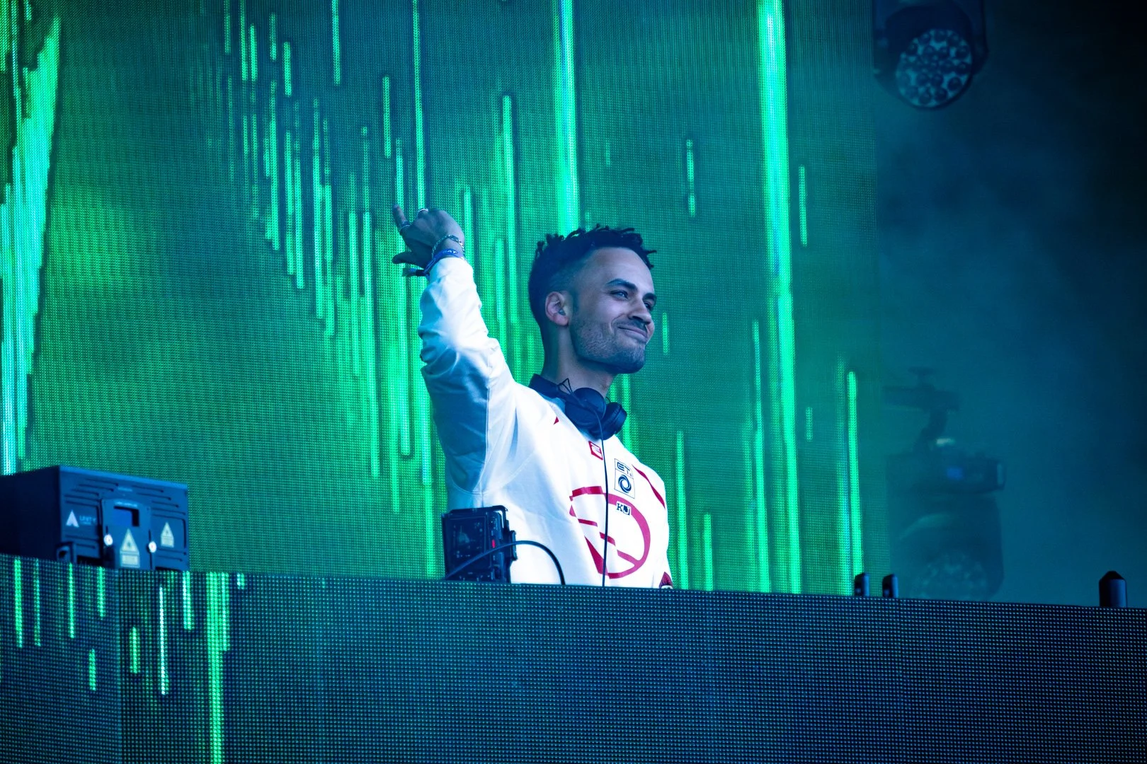 A DJ is standing behind a DJ booth with electronic equipment, wearing a white jacket with red logos, black headphones around his neck, and making a confident expression with a slight smile. The background features a digital green light display.