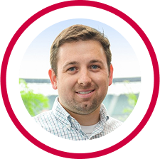Headshot of a smiling man with short brown hair wearing a light blue checked shirt, outdoors with blurred greenery in the background.