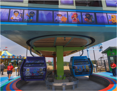 Lego-themed rides at a theme park, including two colorful gondola-style ride vehicles with Lego character scenes, and a backdrop of Lego character posters on windows.