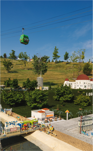 Lego-themed amusement park with mini buildings, pathways, and a gondola lift with a green cabin in the sky, set against a backdrop of trees and a grassy hill with a clear blue sky.