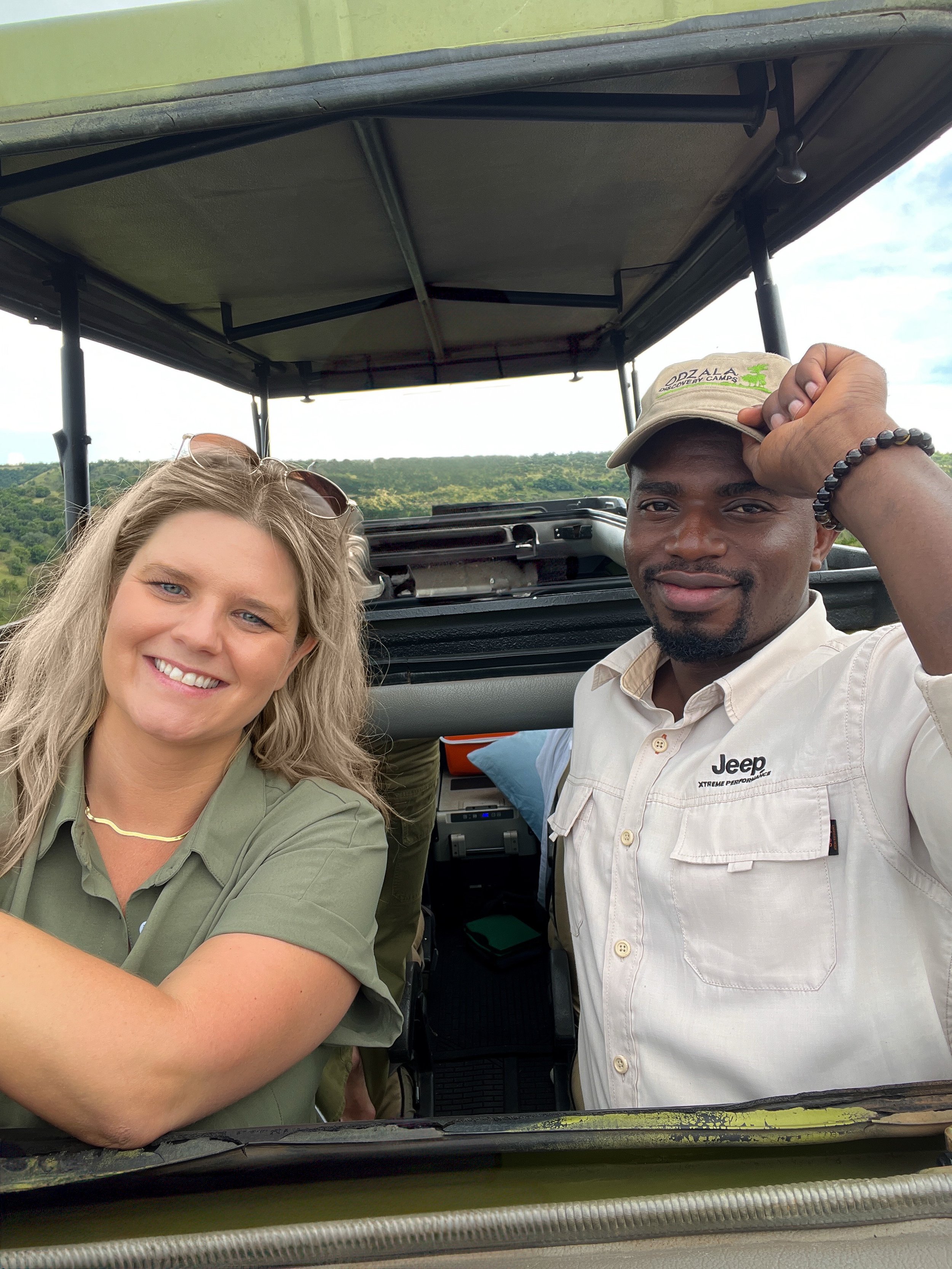 Two people taking a selfie inside a safari vehicle, with a scenic landscape of green hills and cloudy sky in the background.
